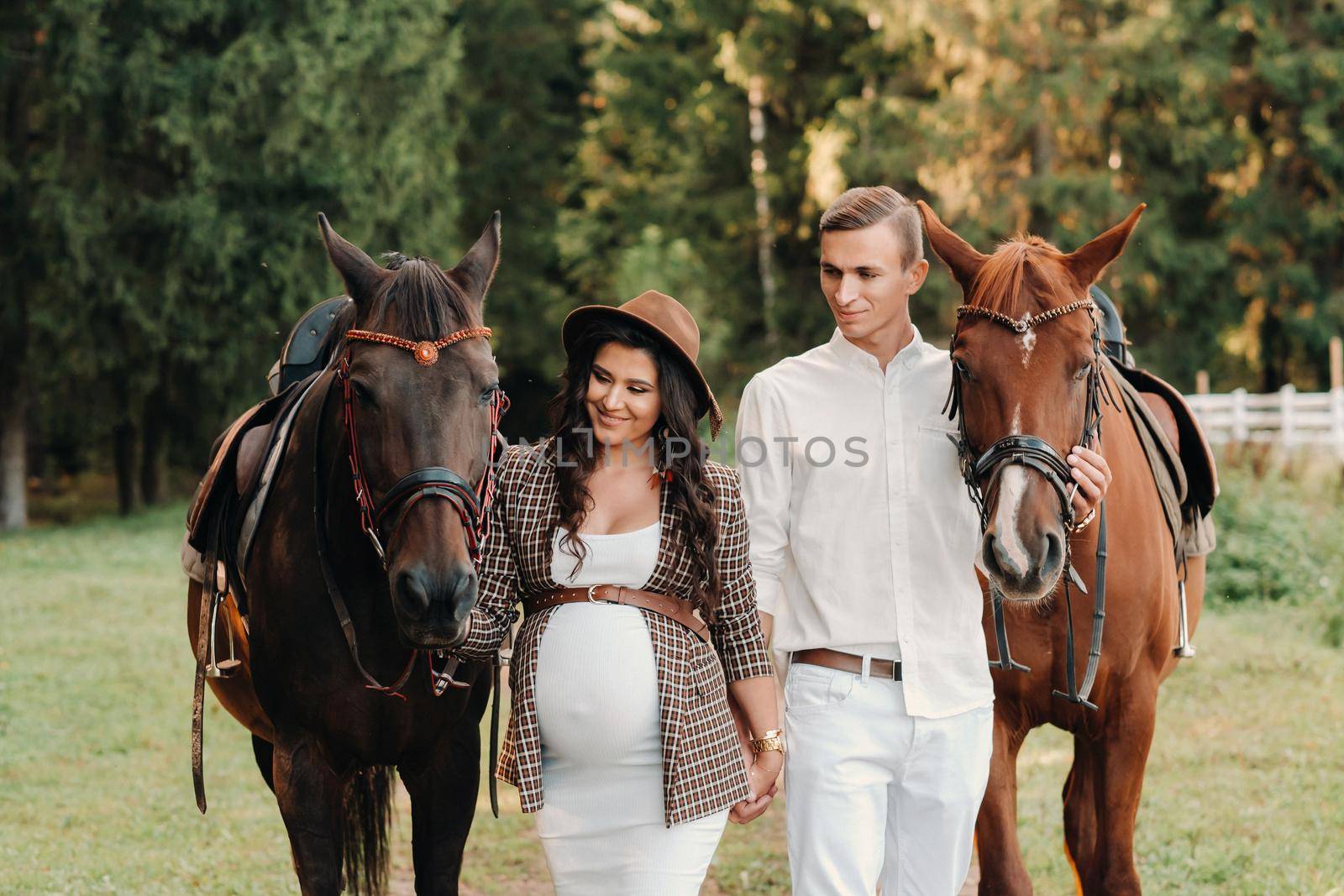 a pregnant girl in a hat and her husband in white clothes stand next to horses in the forest in nature.Stylish pregnant woman with a man with horses.Family