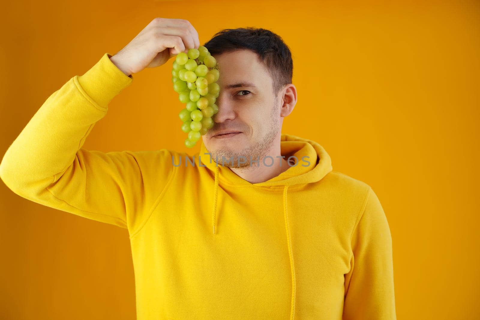 Portrait of young man with green grapes on yellow background. Close up of handsome guy in yellow hoodie closes eye of bunch of grapes