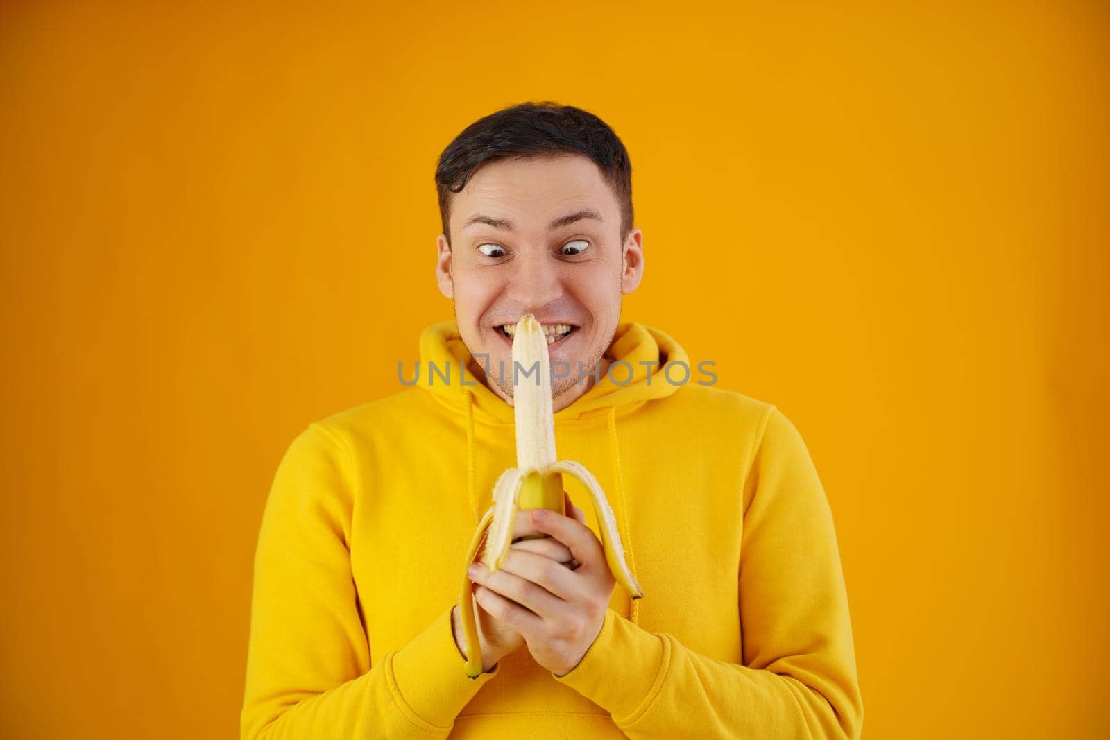 Portrait of young man with banana on yellow background. Funny guy in yellow hoodie posing wth fruit. by epidemiks
