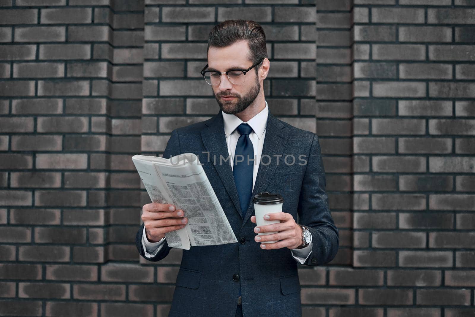 Checking business news. Good looking young man in full suit reading a newspaper on the wall background