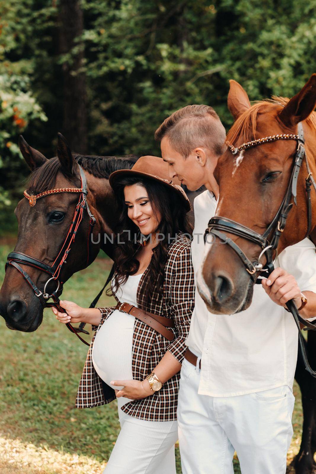 a pregnant girl in a hat and her husband in white clothes stand next to horses in the forest in nature.Stylish pregnant woman with a man with horses.Family