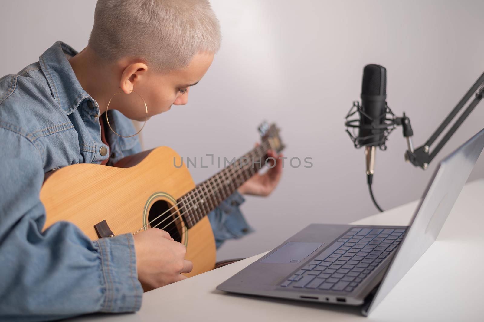 Young caucasian woman with short blonde hair playing guitar and streaming live on laptop