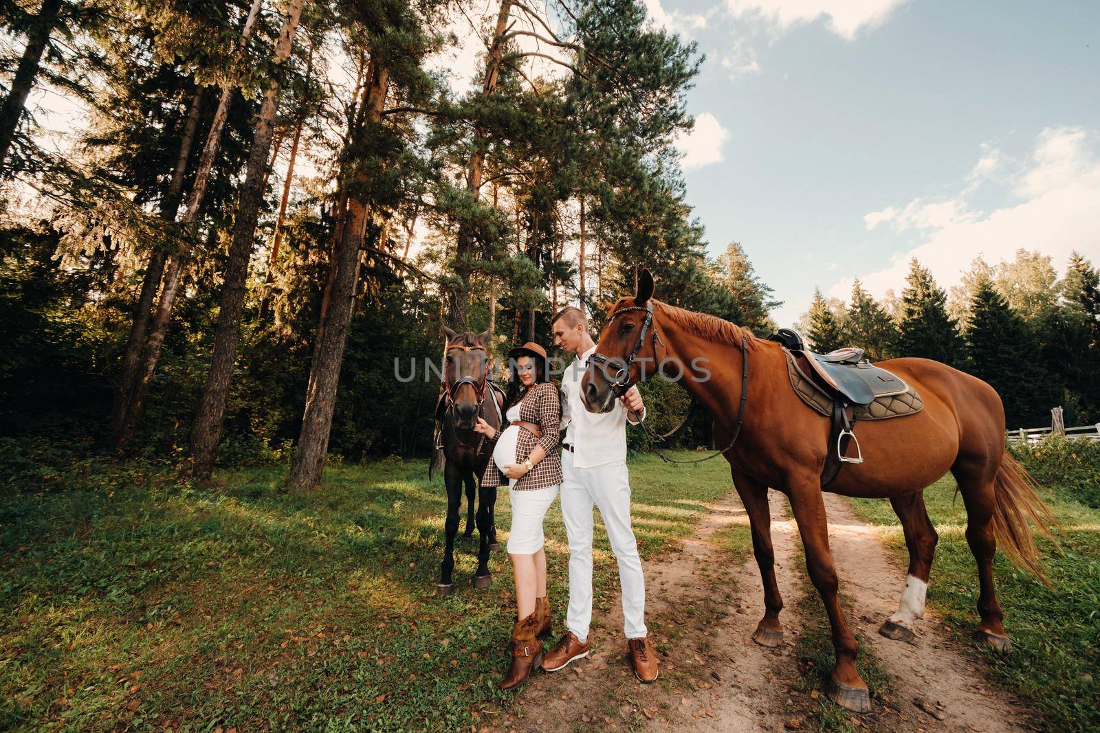 a pregnant girl in a hat and her husband in white clothes stand next to horses in the forest in nature.Stylish pregnant woman with a man with horses.Family