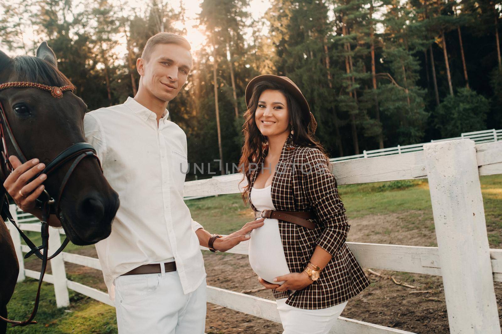 a pregnant girl in a hat and a man in white clothes stand next to horses near a white fence.Stylish pregnant woman with a man with horses.Married couple. by Lobachad