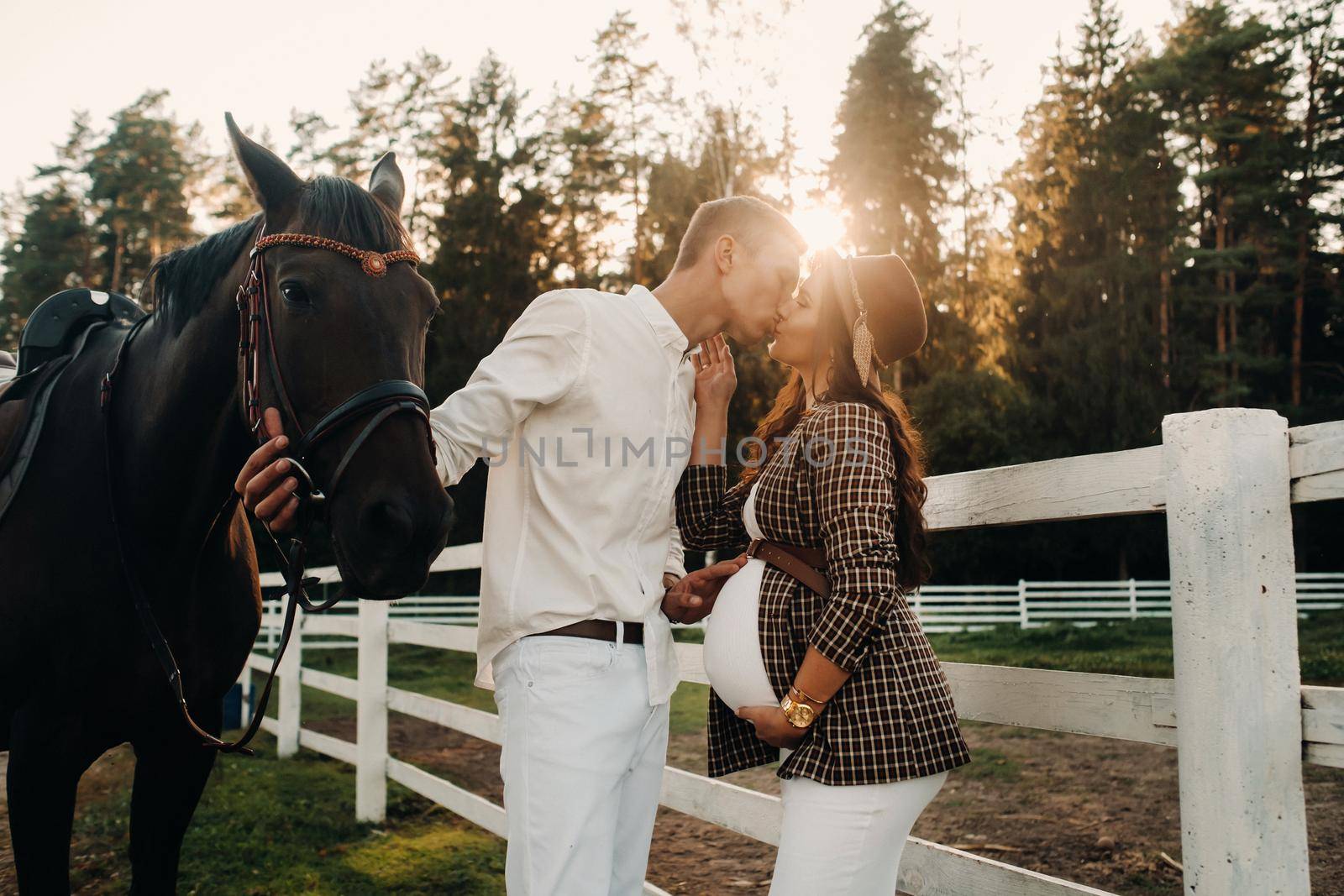 a pregnant girl in a hat and a man in white clothes stand next to horses near a white fence.Stylish pregnant woman with a man with horses.Married couple. by Lobachad