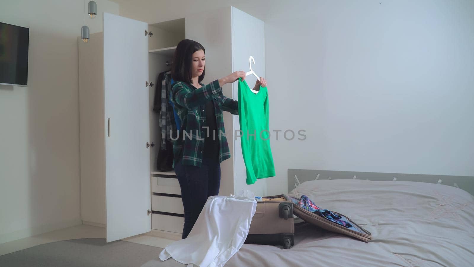 Young brunette Hanging clothes in a wardrobe. On the bed suitcase with female dress. Girl wearing in casual jeans and shirt in small flat with white interior.