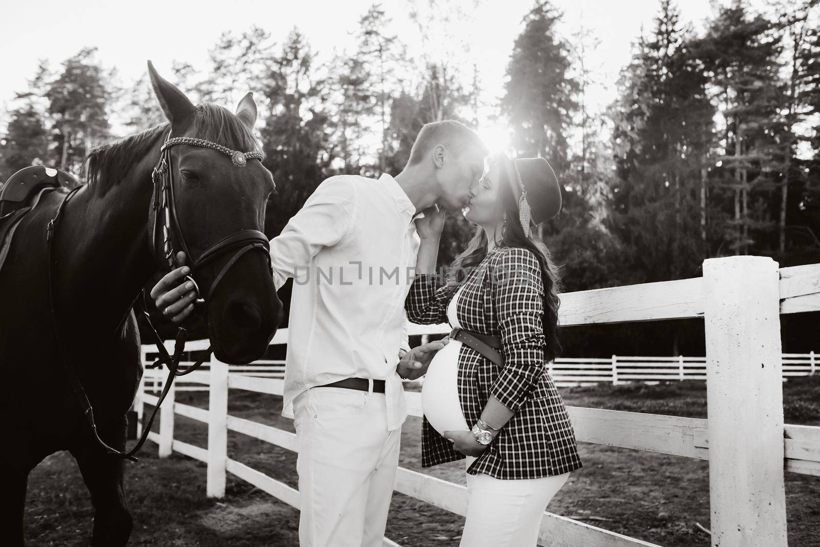 a pregnant girl in a hat and her husband in white clothes stand next to the horses near the horse corral at sunset.Stylish pregnant woman with a man with horses.Family. black and white photo.