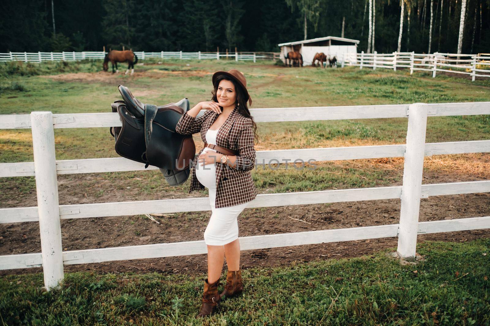 a pregnant girl with a big belly in a hat next to horses near a paddock in nature.Stylish pregnant woman in a brown dress with horses. by Lobachad