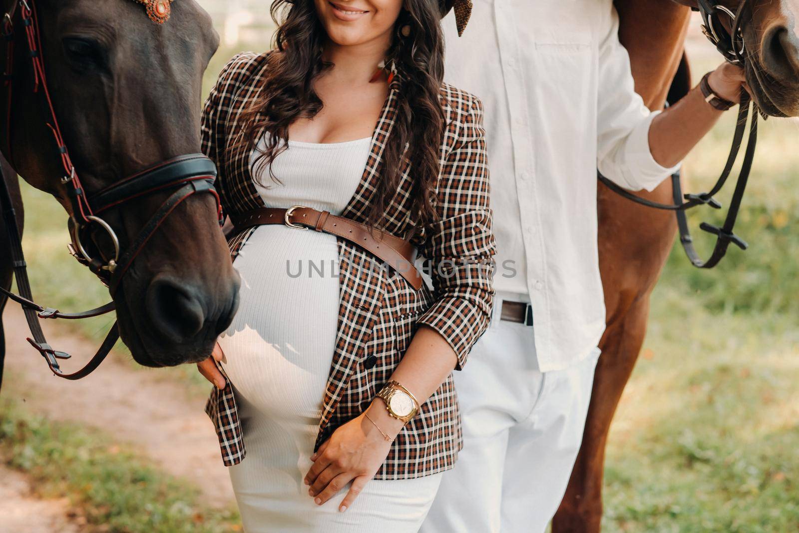 a pregnant girl in a hat and her husband in white clothes stand next to horses in the forest in nature.Stylish pregnant woman with a man with horses.Family