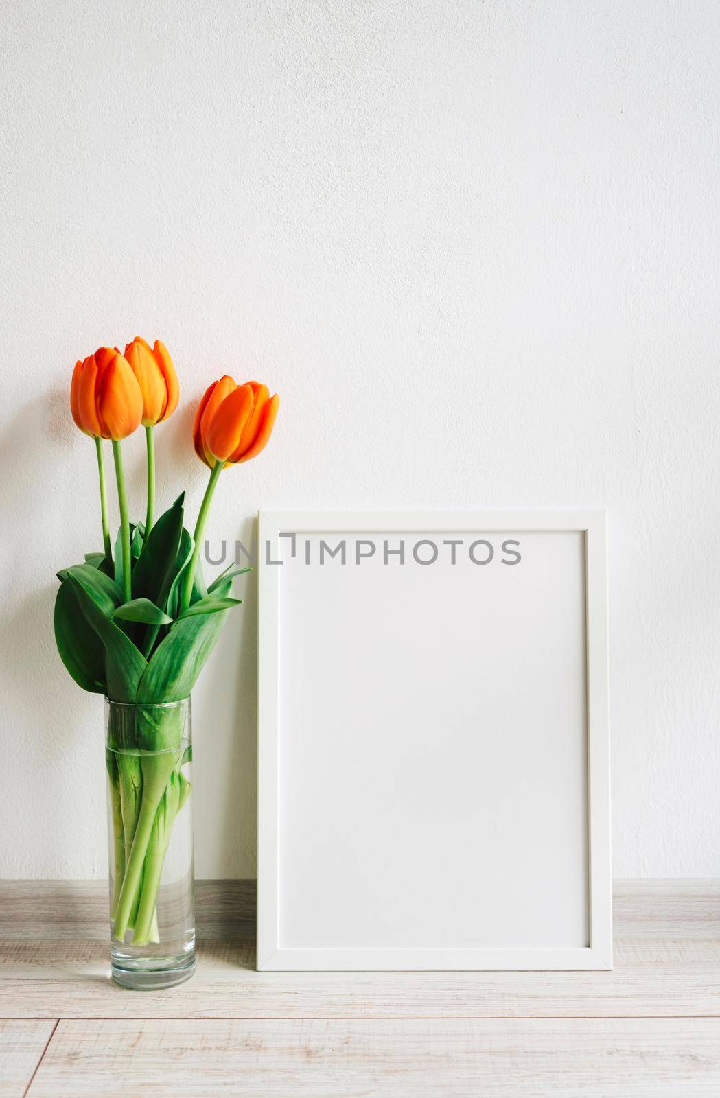 Poster frame mockup. White wooden frame for painting, photo or poster. Bright orange bouquet of fresh tulips in a glass vase.
