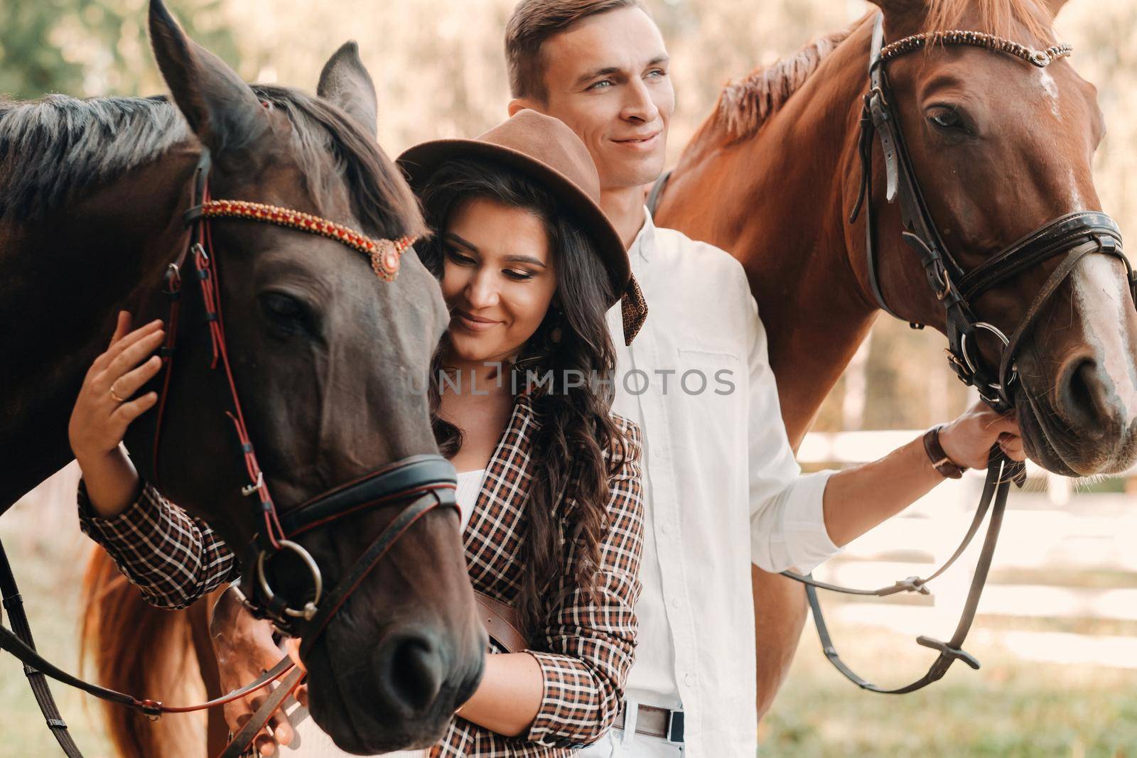 a pregnant girl in a hat and her husband in white clothes stand next to horses in the forest in nature.Stylish pregnant woman with a man with horses.Family