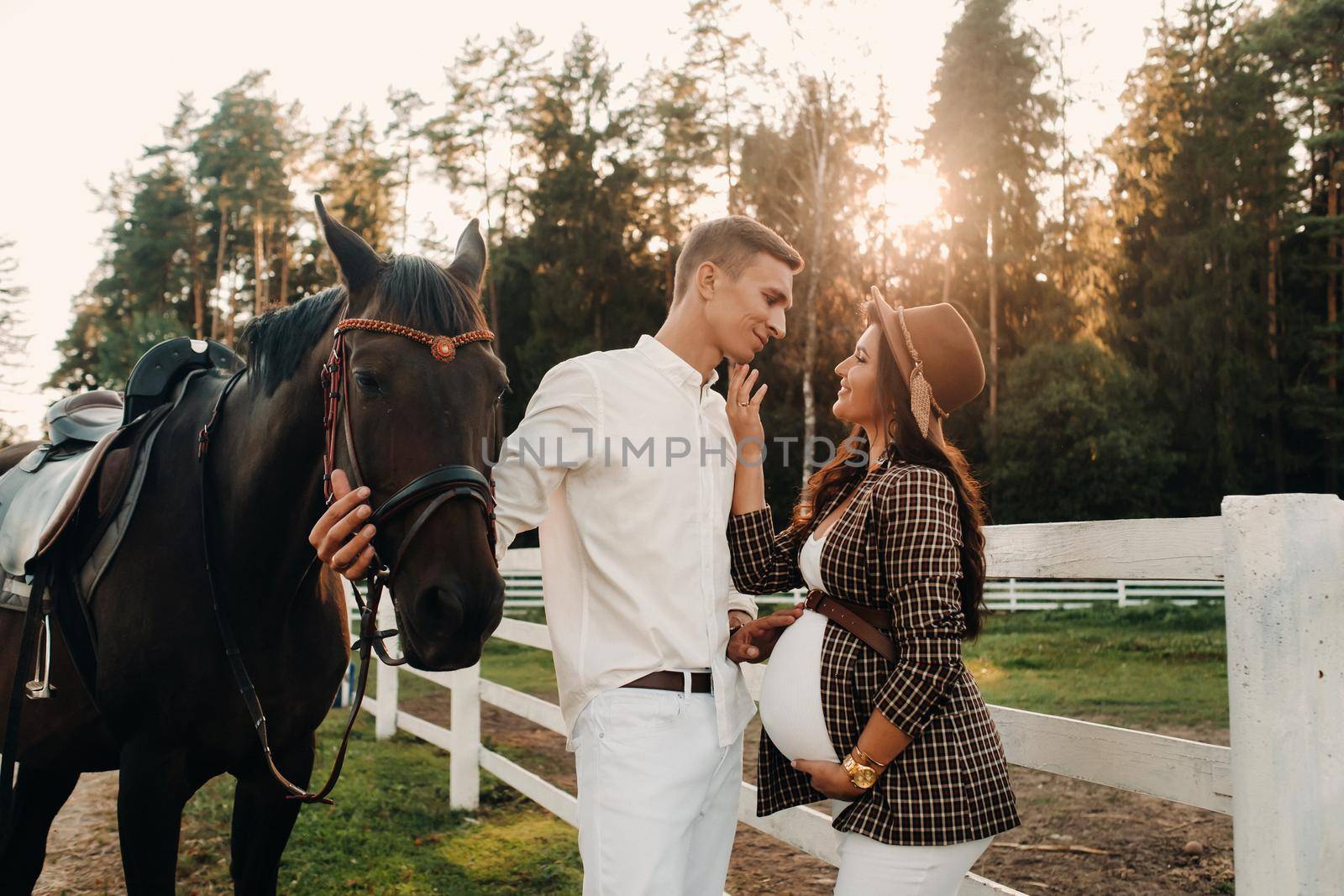 a pregnant girl in a hat and a man in white clothes stand next to horses at a White fence.Stylish family waiting for a child strolling in nature by Lobachad