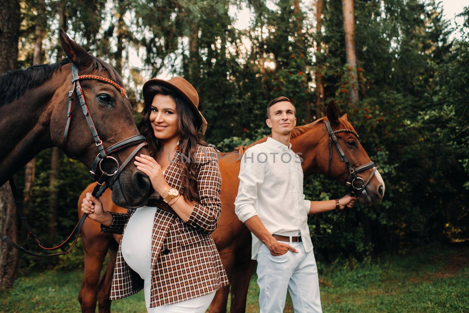 a pregnant girl in a hat and a man in white clothes stand next to horses in the forest in nature.Stylish pregnant woman with her husband with horses.Married couple
