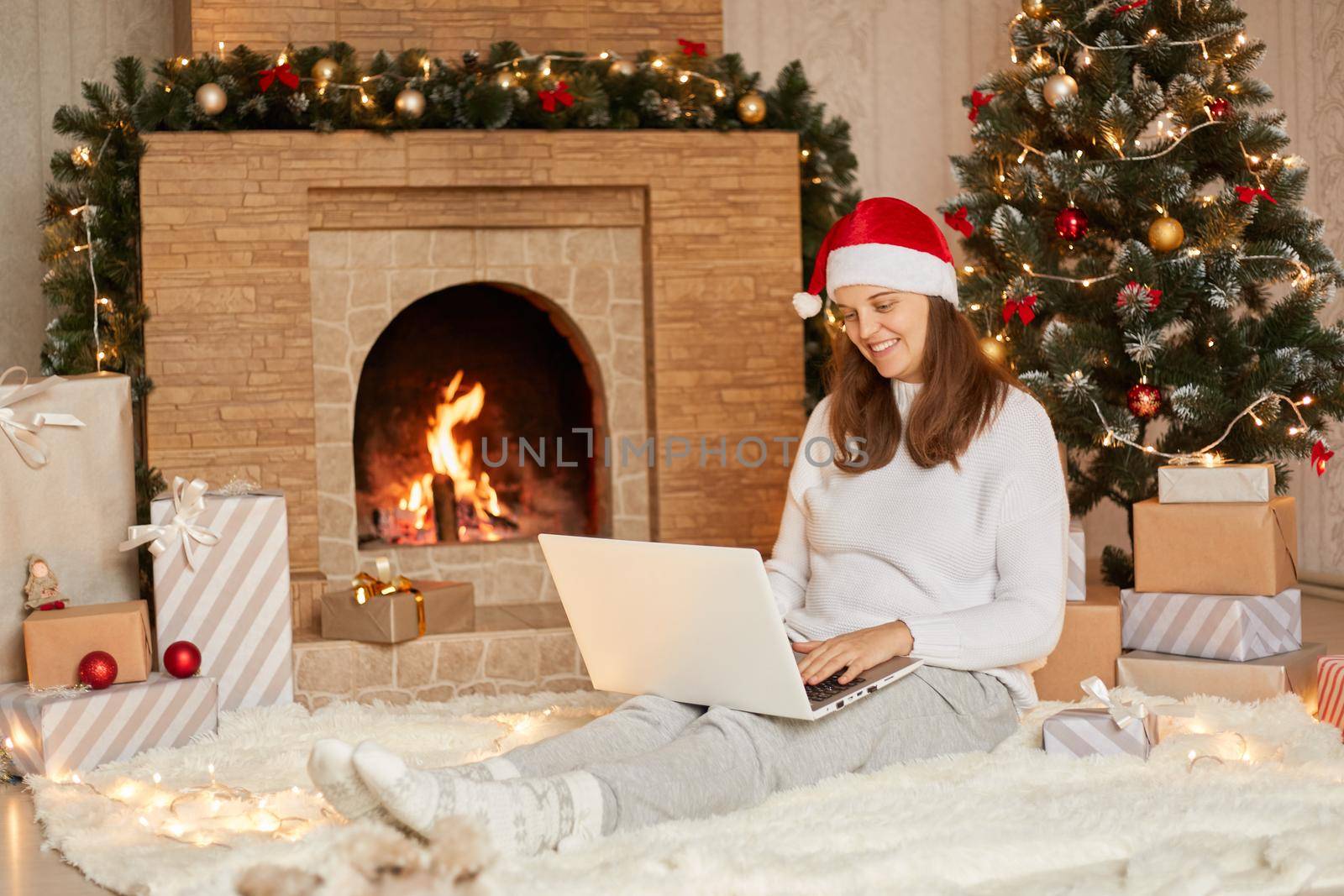 Photo of cute nice charming attractive girl having video call in eve of new year with her laptop, wearing casual white sweater and red hat, looks at device's camera with charming smile. by sementsovalesia