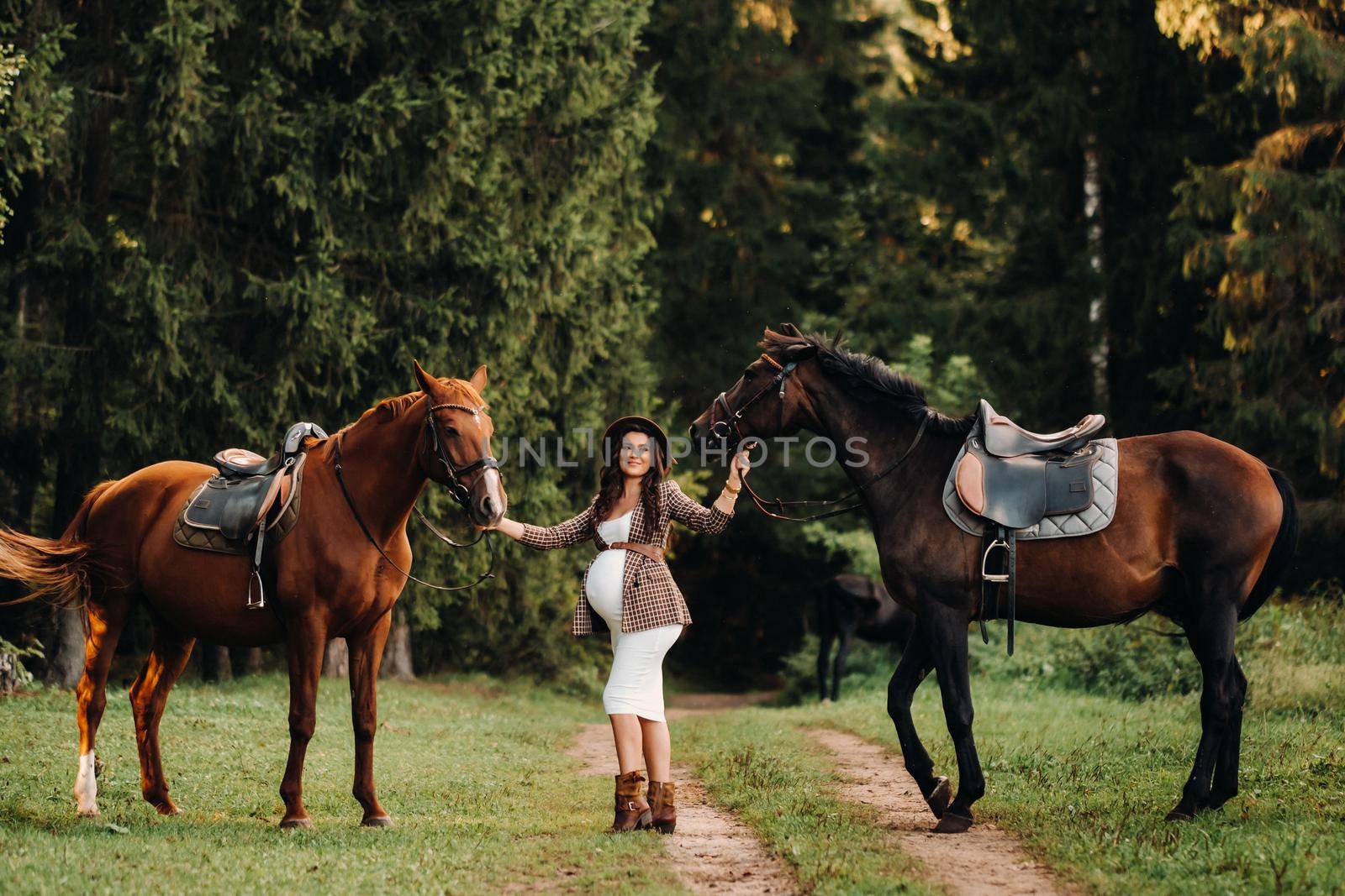 pregnant girl with a big belly in a hat next to horses in the forest in nature.Stylish pregnant woman in the brown dress with the horses