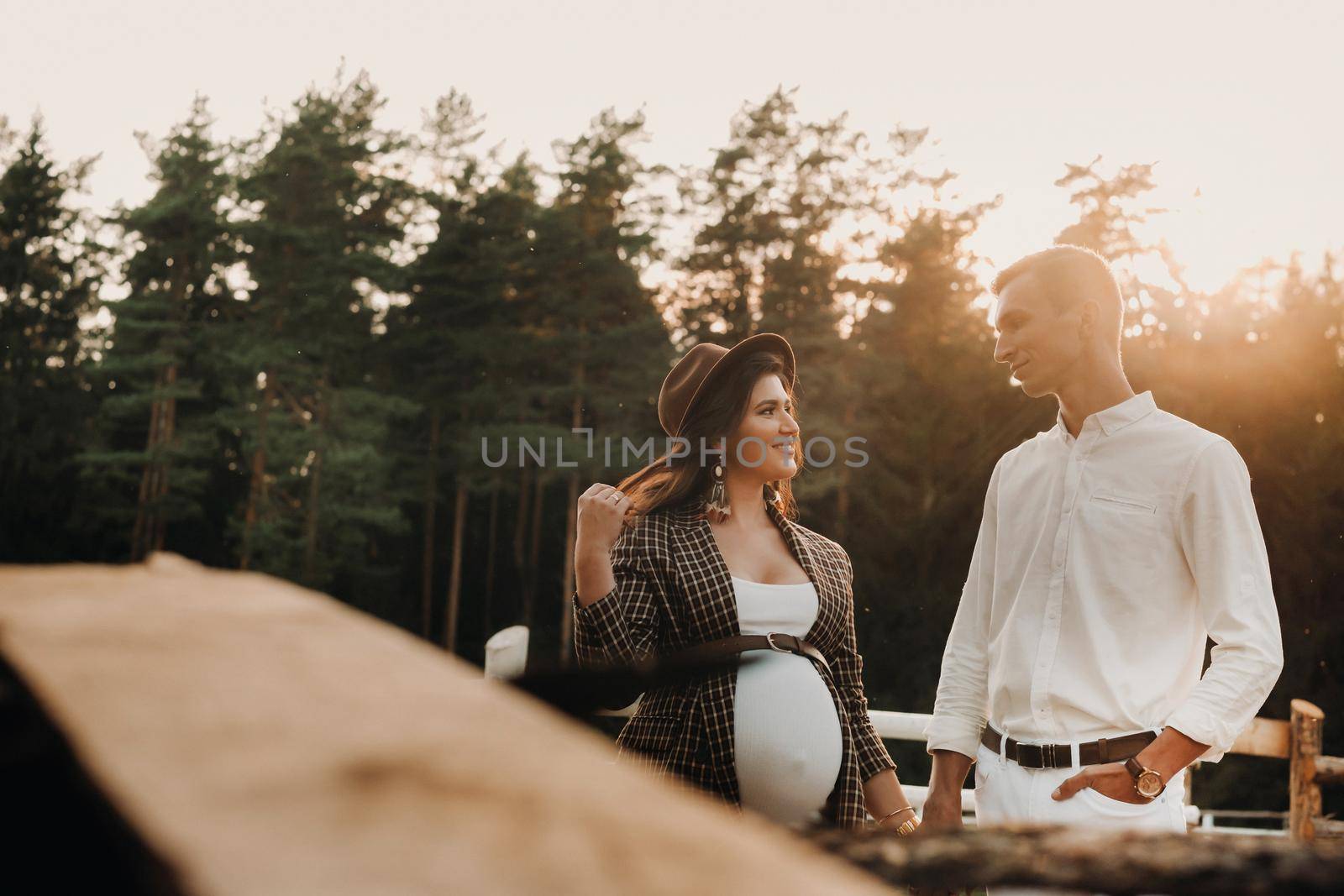 a pregnant girl in a hat and her husband in white clothes stand next to a horse corral at sunset.a stylish couple is waiting for a child in nature.