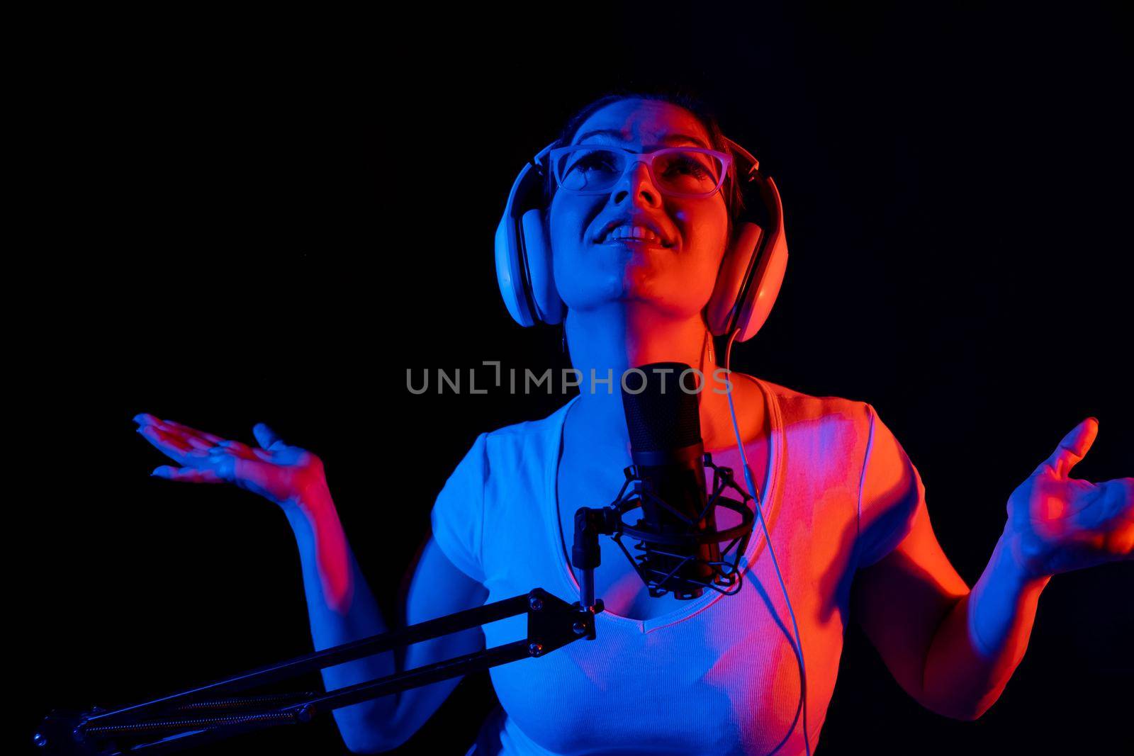 Caucasian woman in glasses and headphones sings into a microphone in neon light on a black background. An emotional girl is recording a song in a recording studio.