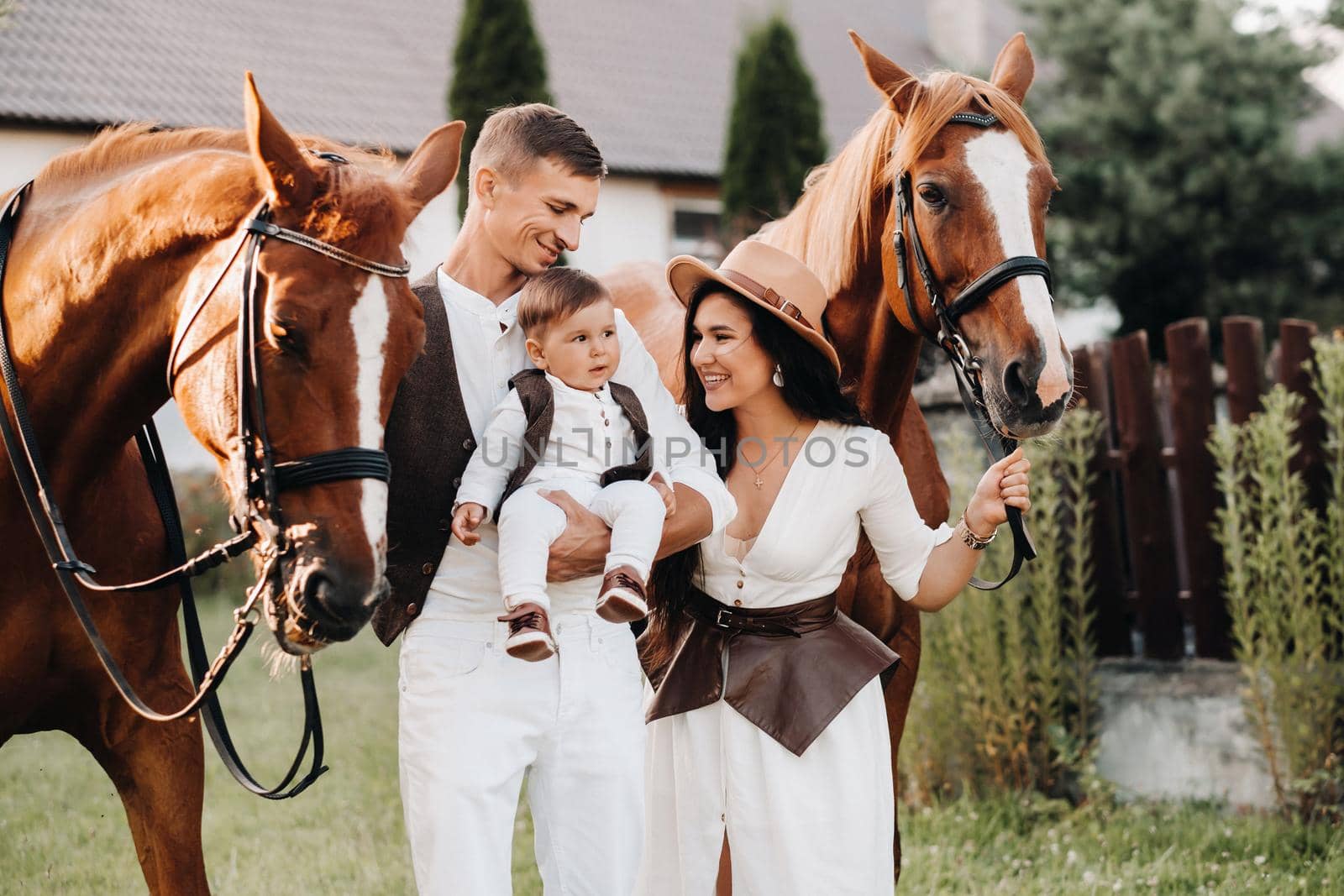 A family in white clothes with their son stand near two beautiful horses in nature. A stylish couple with a child are photographed with horses by Lobachad