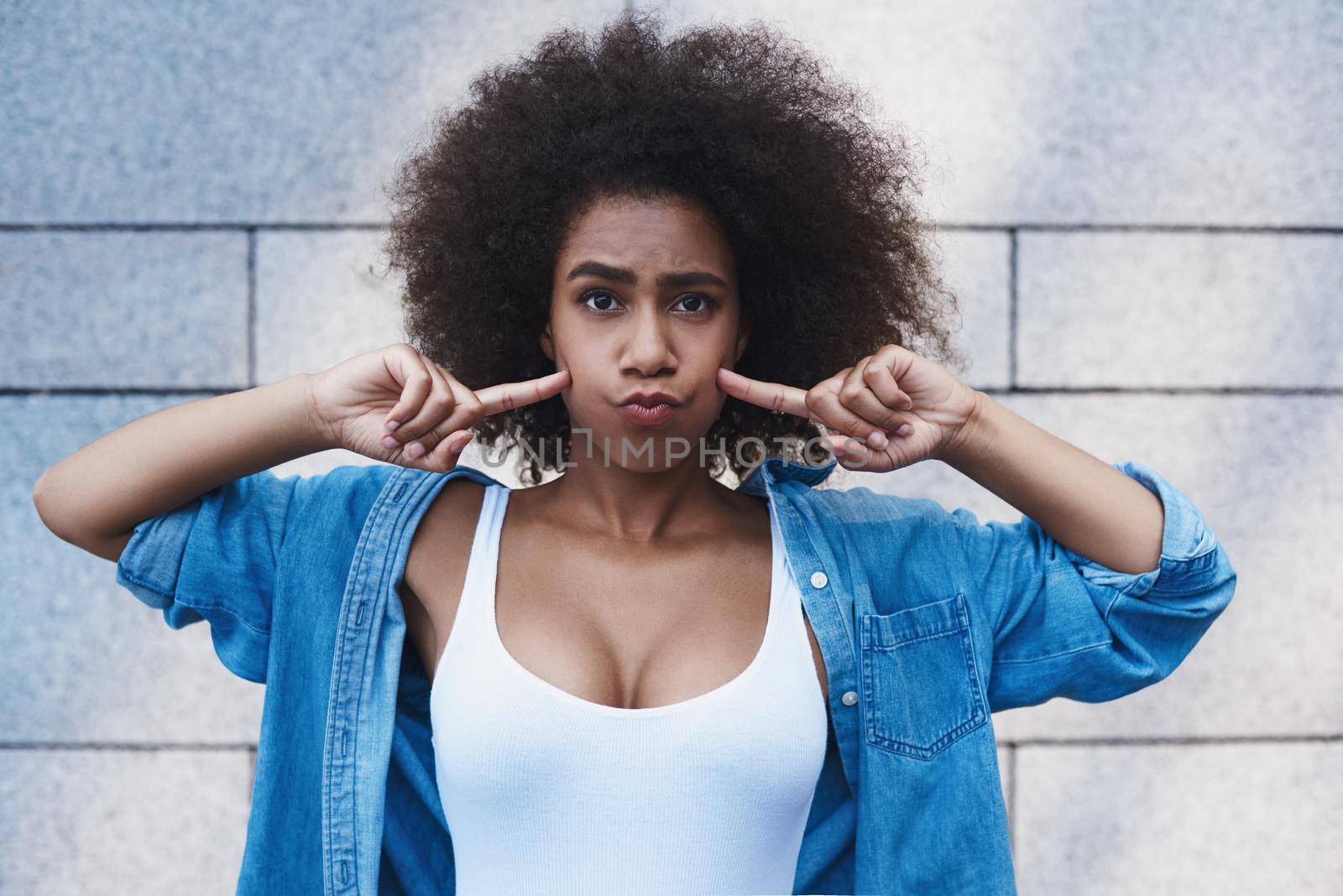 Young woman wearing jeans jacket free style on the street standing isolated on concrete wall touching cheeks pouting lips childish