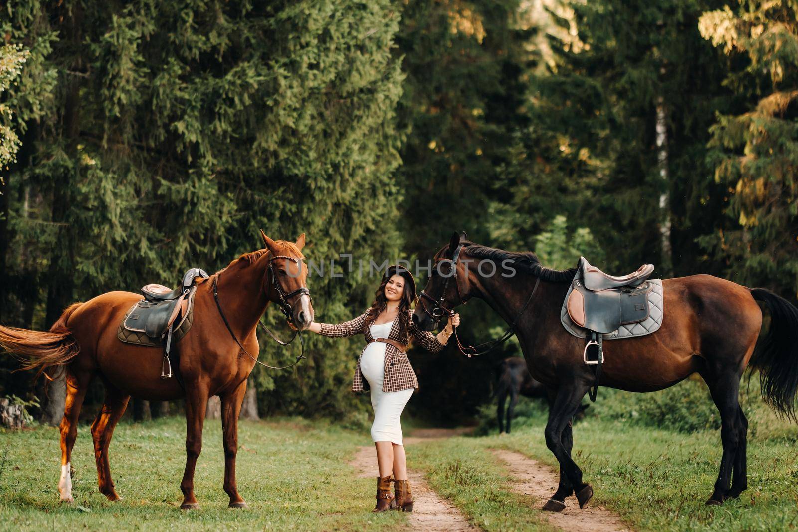 pregnant girl with a big belly in a hat next to horses in the forest in nature.Stylish girl in white clothes and a brown jacket