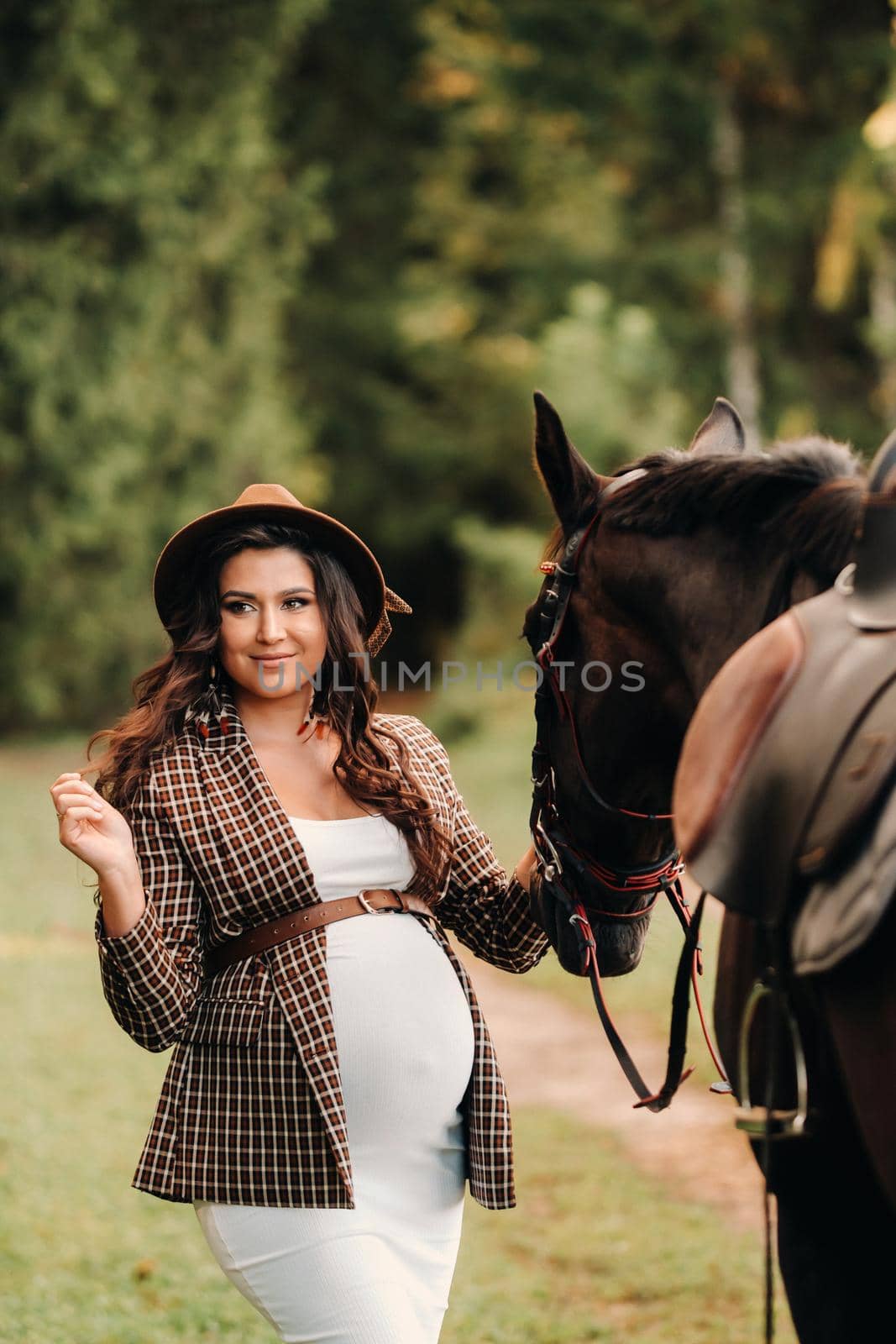 pregnant girl with a big belly in a hat next to horses in the forest in nature.A stylish pregnant woman in a white dress and brown jacket with horses