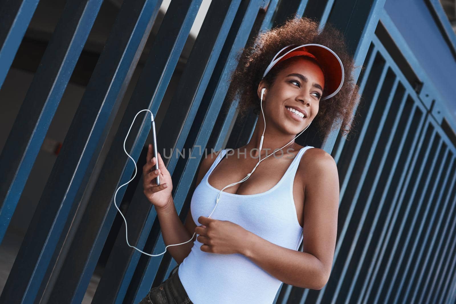Young woman wearing tennis visor and earphones free style on the street standing near fence holding smartphone listening music looking aside smiling happy
