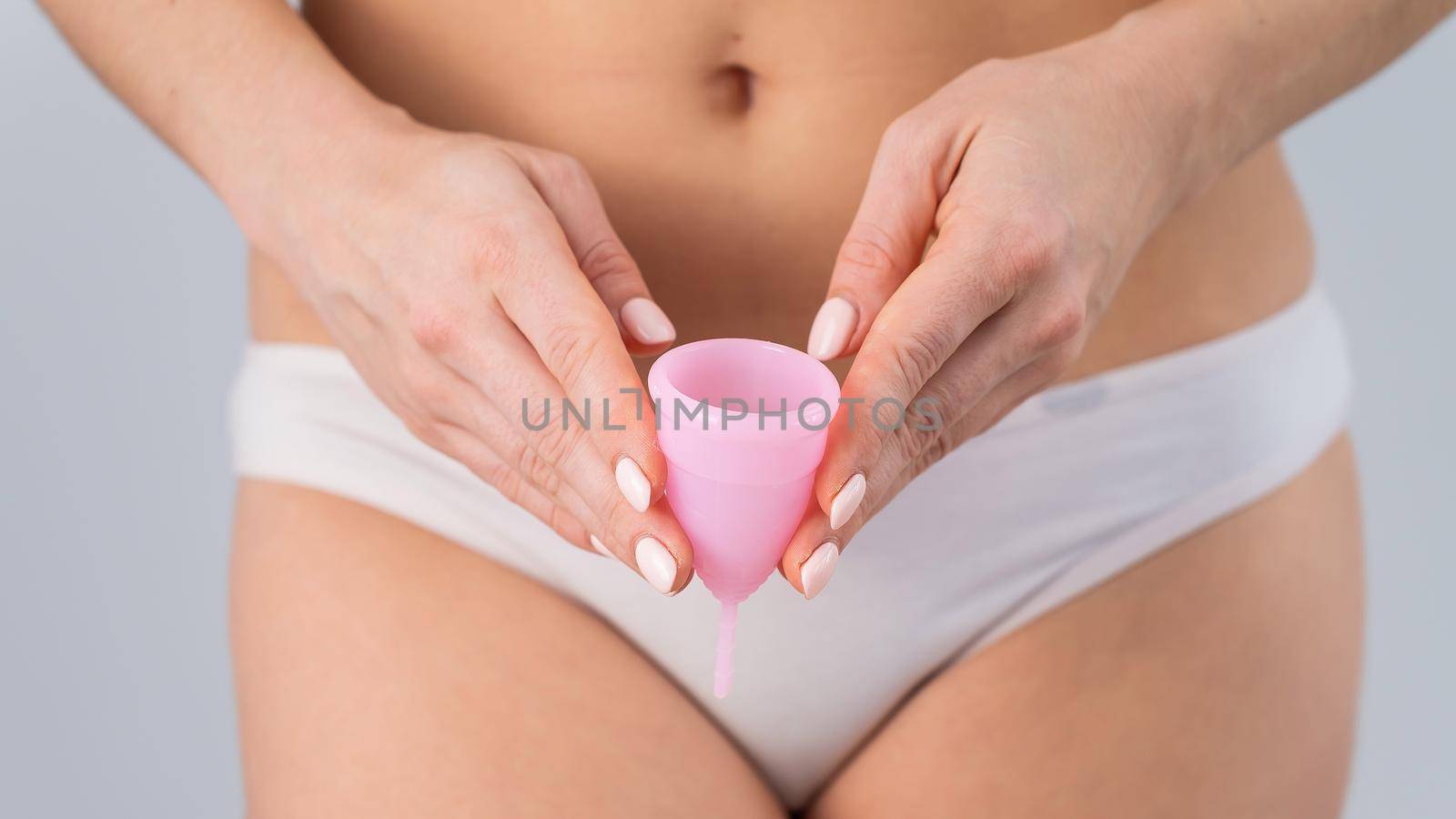 Close-up of a woman in white cotton panties holding a pink menstrual cup against a white background. Alternative to tampons and pads.