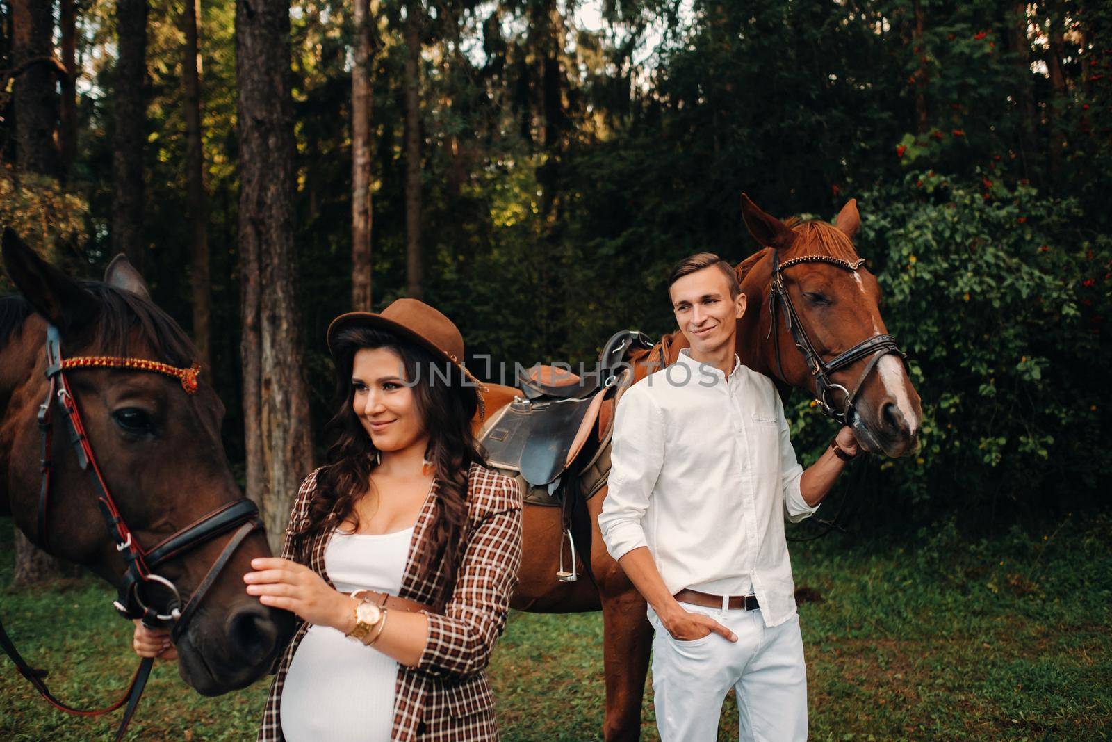 a pregnant girl in a hat and a man in white clothes stand next to horses in the forest in nature.Stylish pregnant woman with her husband with horses.Married couple
