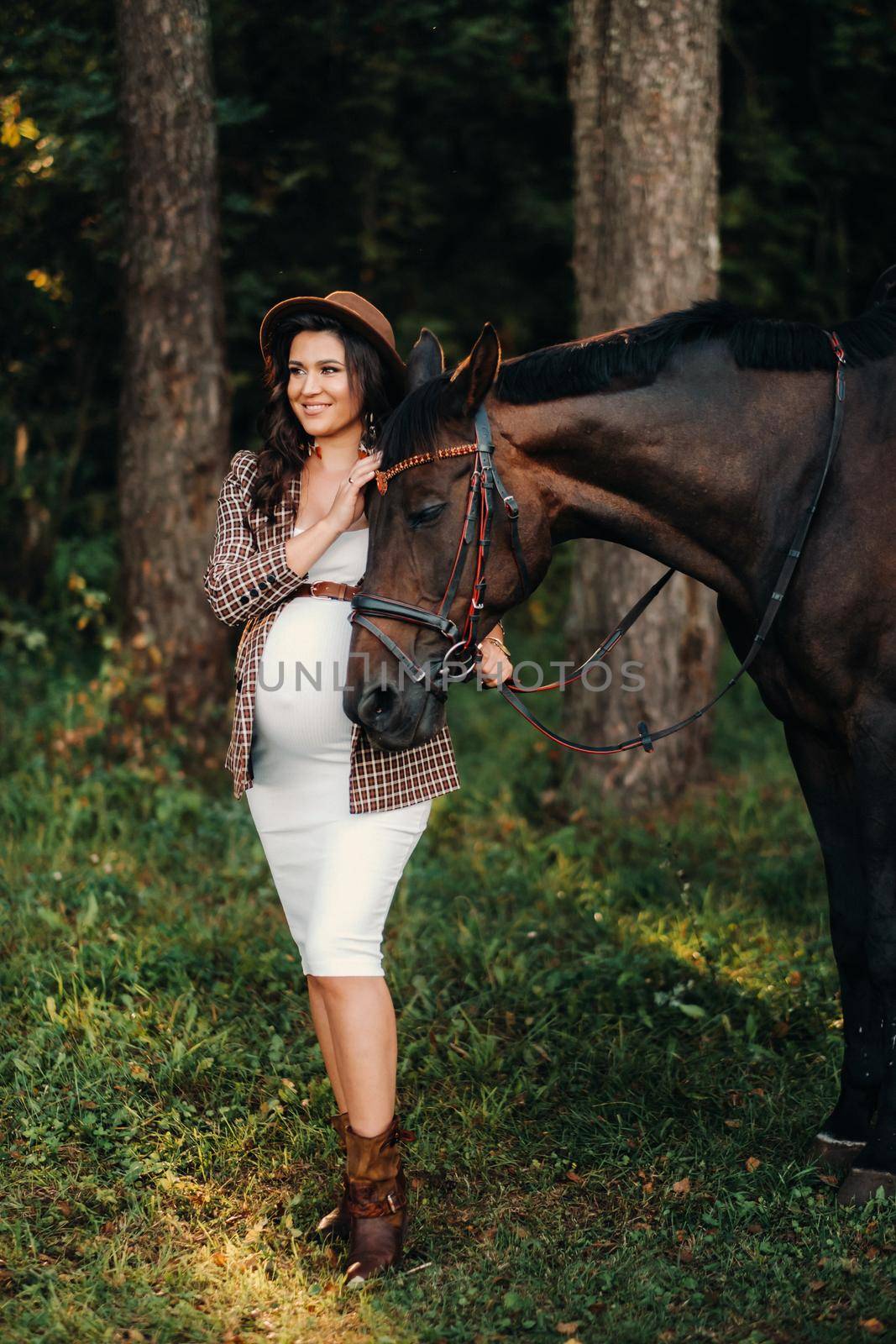 pregnant girl with a big belly in a hat next to horses in the forest in nature.Stylish girl in white clothes and a brown jacket