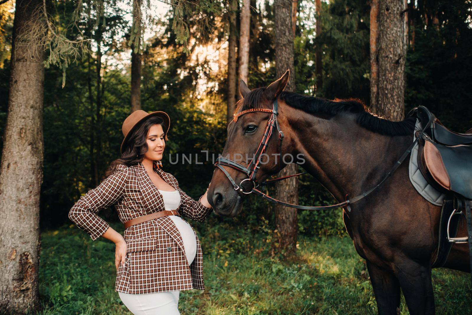 pregnant girl with a big belly in a hat next to horses in the forest in nature.Stylish girl in white clothes and a brown jacket