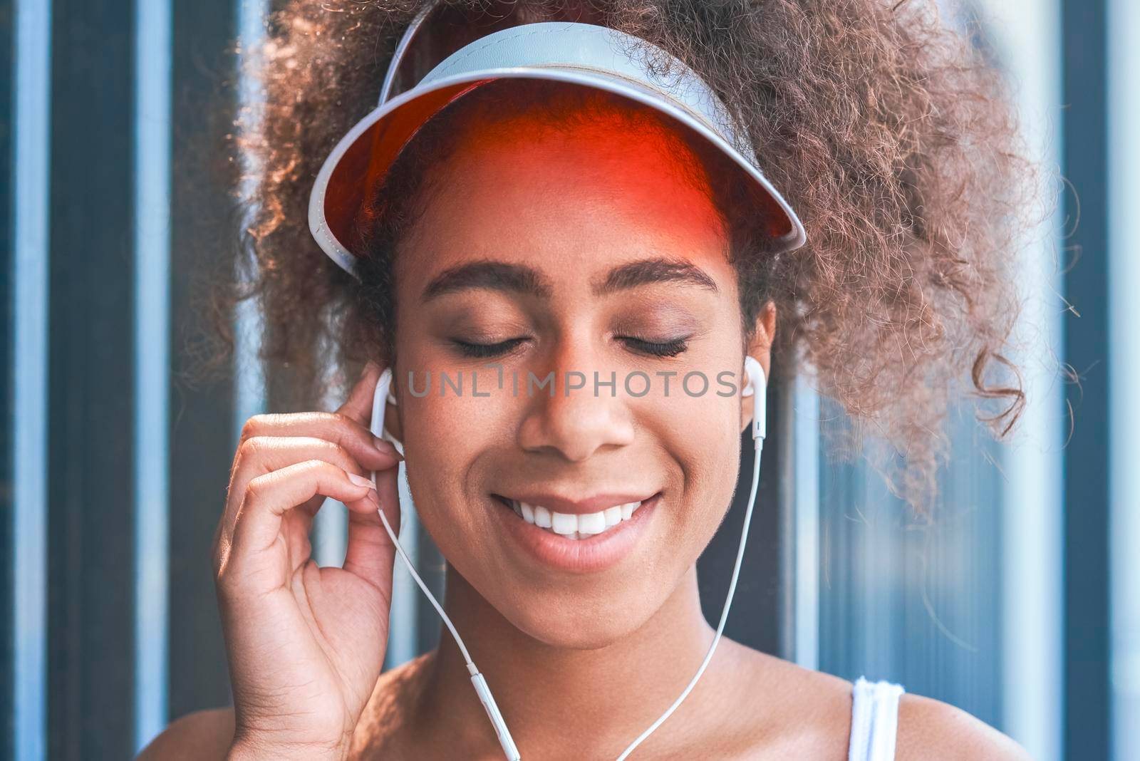 Young woman wearing tennis visor putting on earphones free style on the street standing near fence closed eyes smiling happy close-up