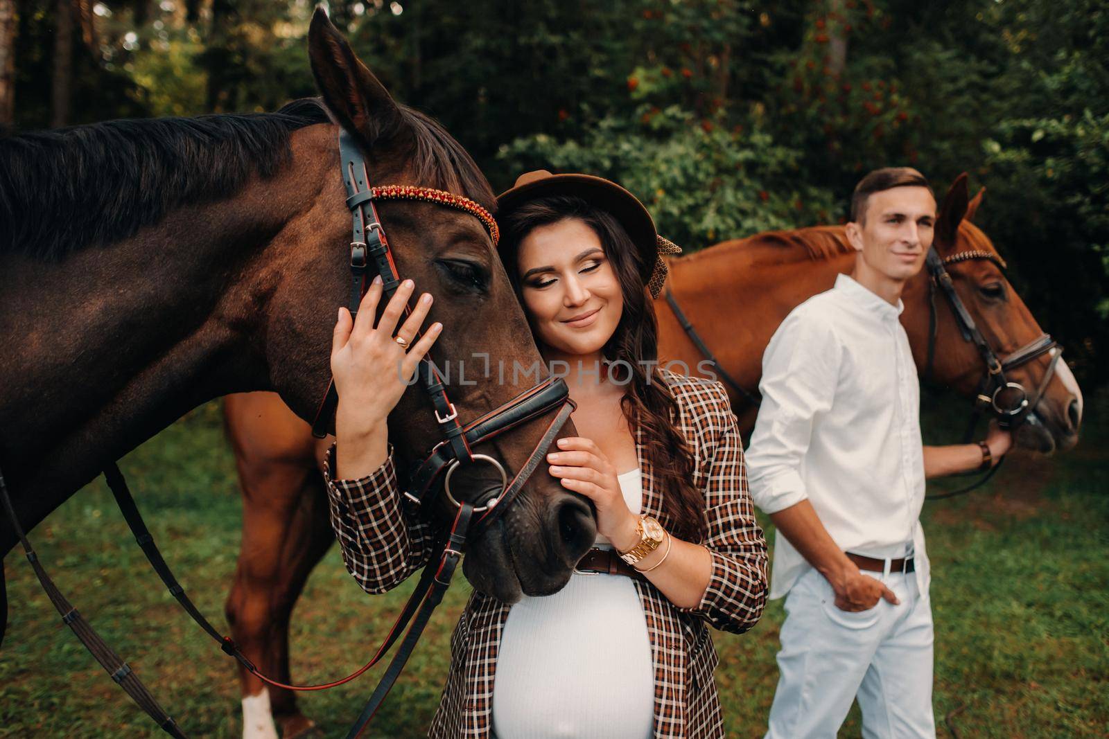 a pregnant girl in a hat and a man in white clothes stand next to horses in the forest in nature.Stylish pregnant woman with her husband with horses.Married couple. by Lobachad