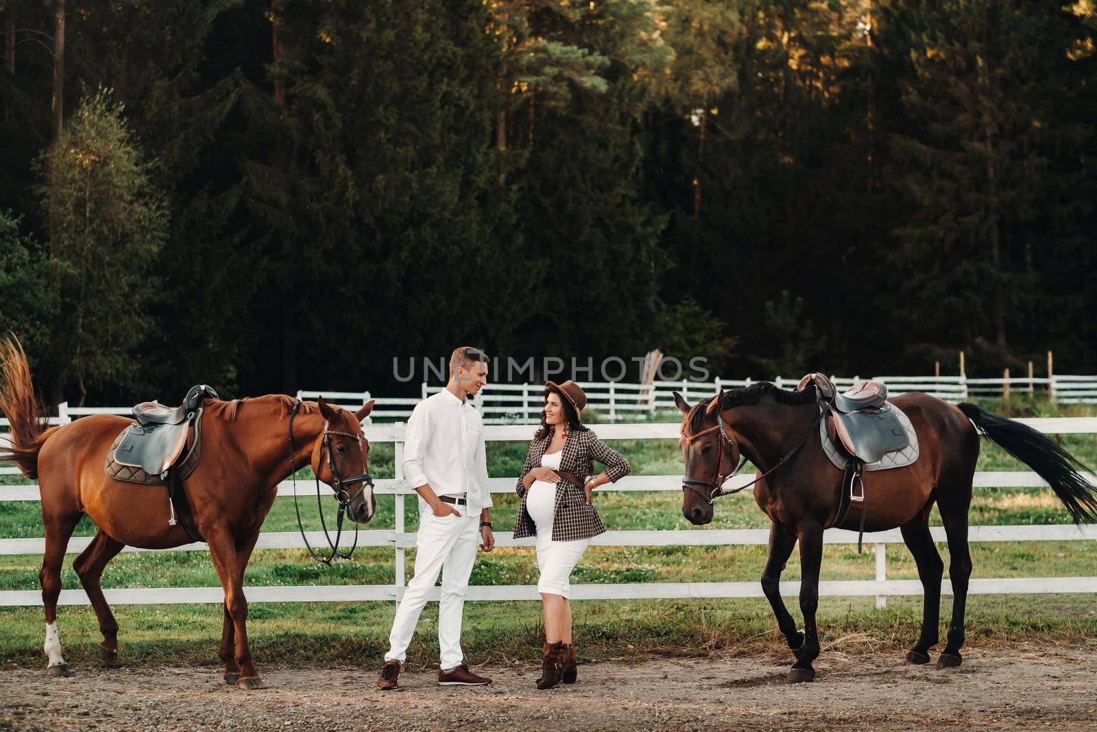 a pregnant girl in a hat and a man in white clothes stand next to horses near a white fence.Stylish pregnant woman with a man with horses.Married couple. by Lobachad
