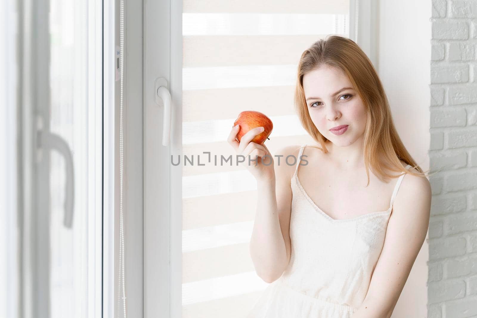 beautiful young woman in a white dress holding a red apple by the window in a bright room