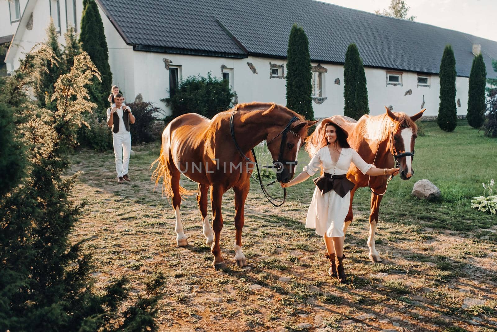 A family in white clothes with their son walking with two beautiful horses in nature. A stylish couple with a child are photographed with horses at sunset by Lobachad