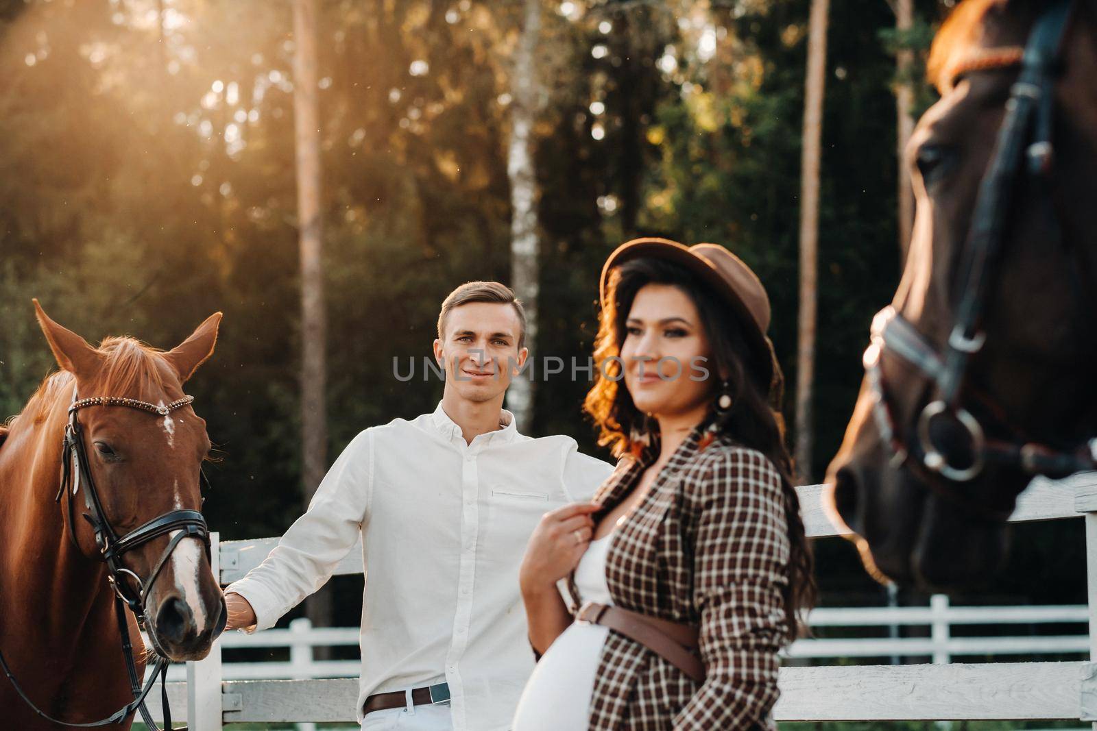 a pregnant girl in a hat and her husband in white clothes stand next to the horses near the horse corral.Stylish pregnant woman with a man with horses.Family.