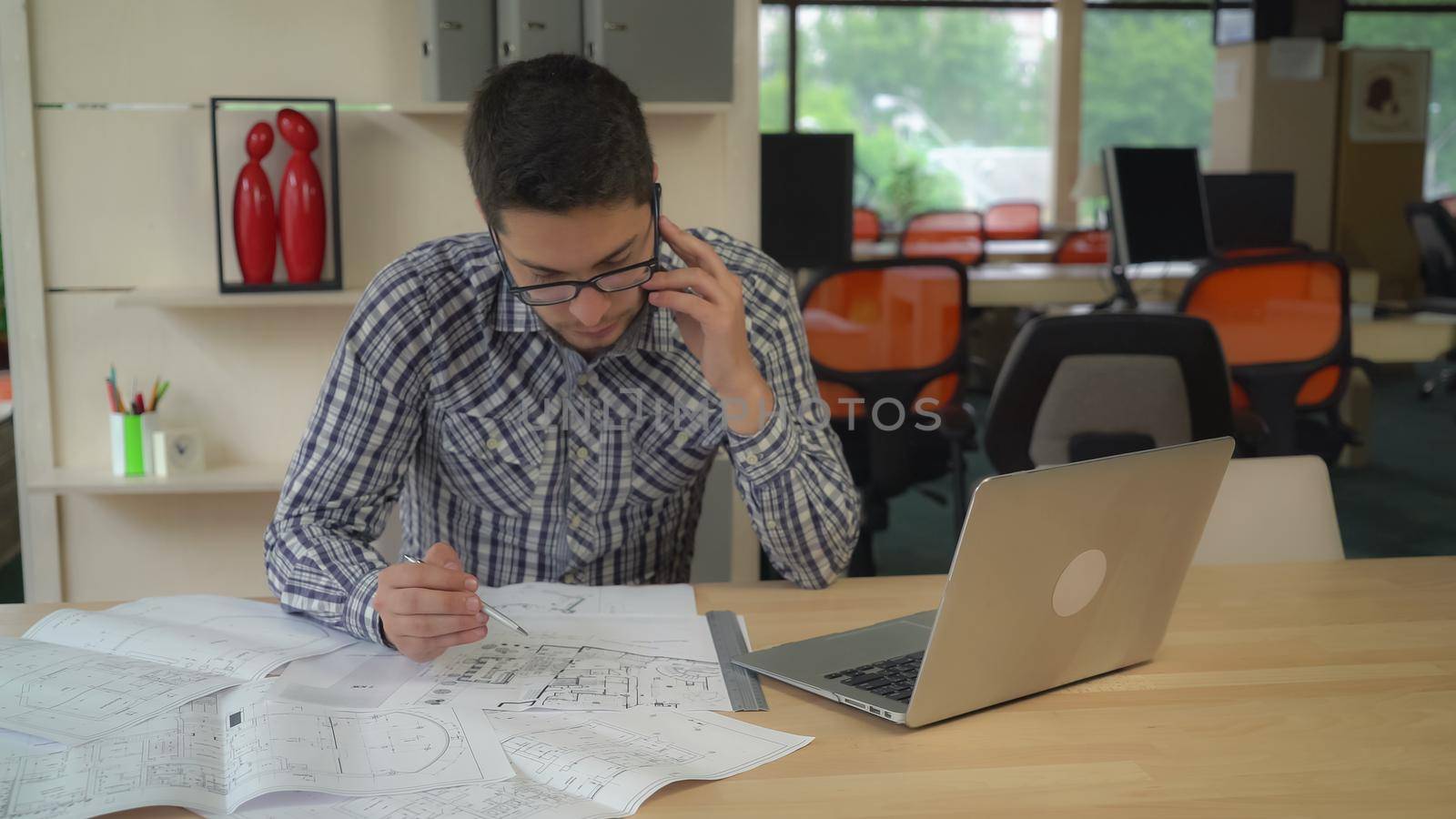 Architect checking details and make notes on the paper plan. Blueprint documents and computer on the wooden table with equipment for drawing and draftsmanship. Male wearing in casual clothes and glasses with black frame work in modern creative office with big windows.