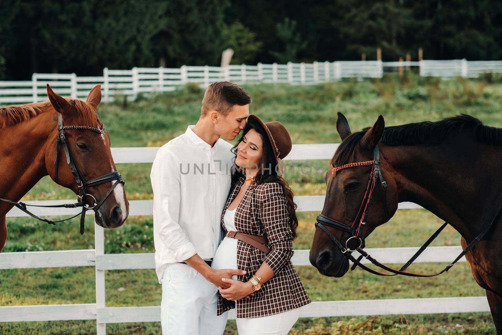 a pregnant girl in a hat and her husband in white clothes stand next to the horses near the horse corral.Stylish pregnant woman with a man with horses.Family by Lobachad