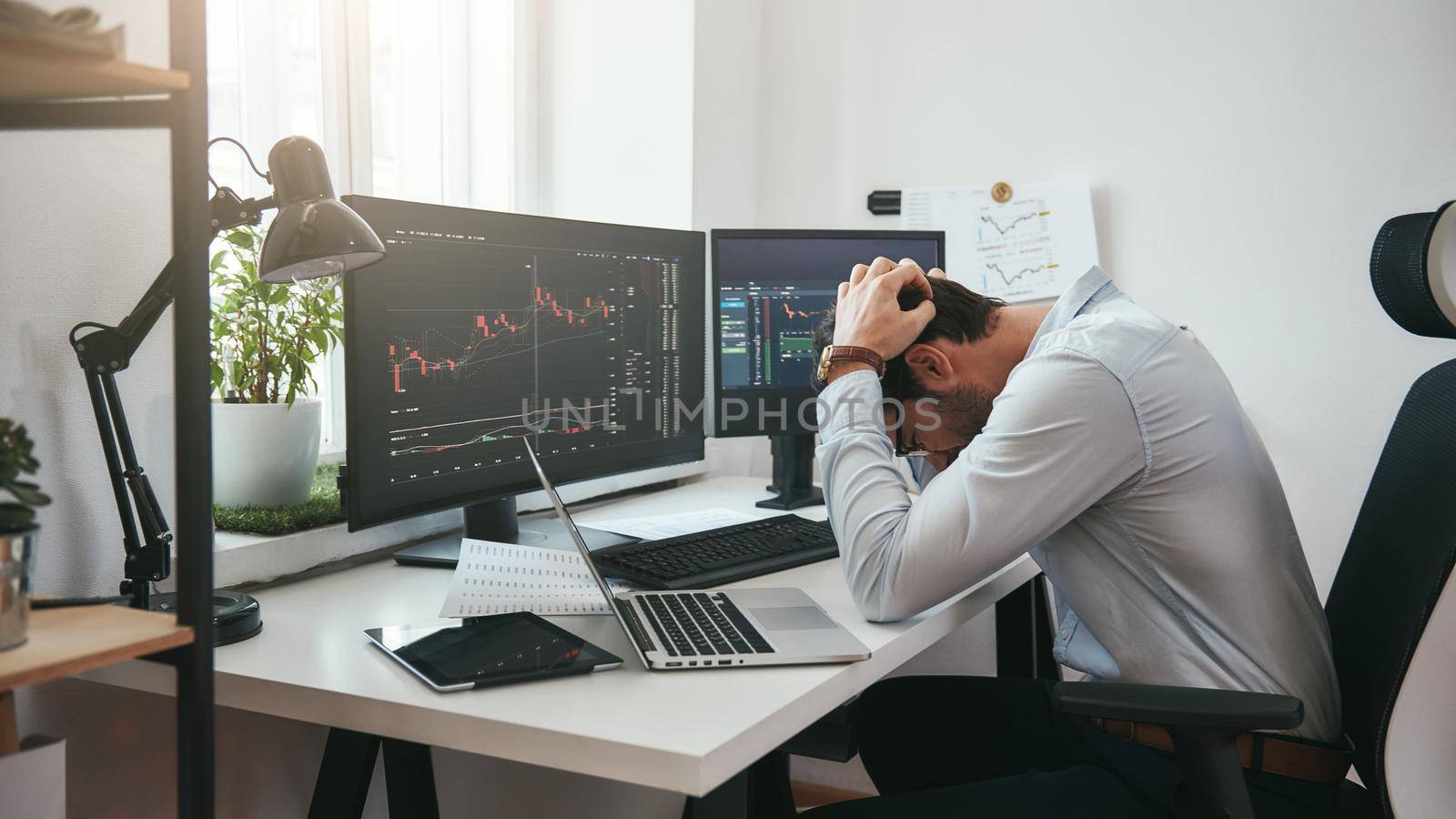 Feeling sad. Depressed young businessman or trader in formalwear keeping head in hands while working in the office. Computer screens with trading charts and financial data. by friendsstock