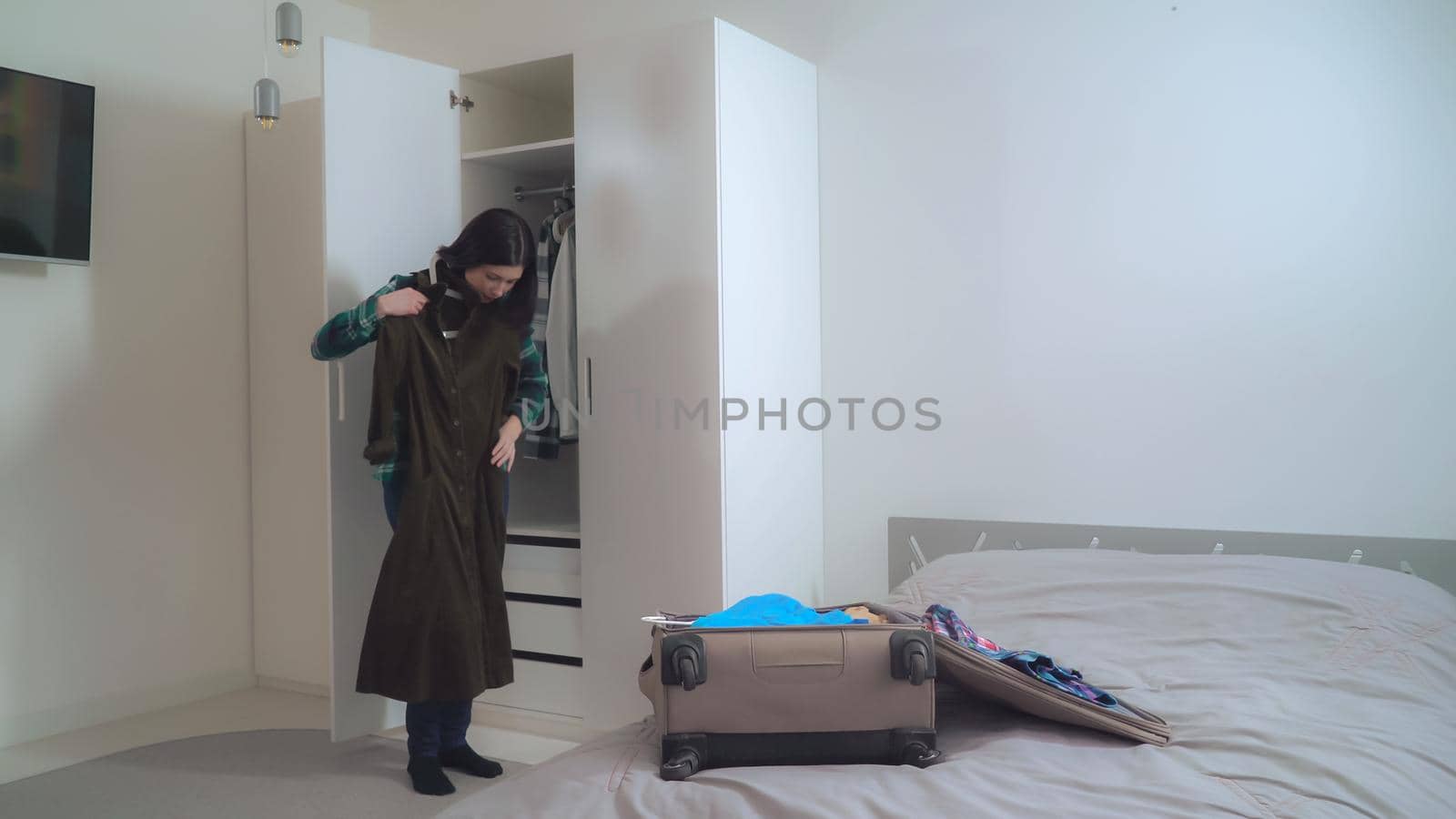 Lady try on dress and put it in suitcase. Young woman wearing in casual clothes packing bag planning vocation. Caucasian girl with black hair in apartment.