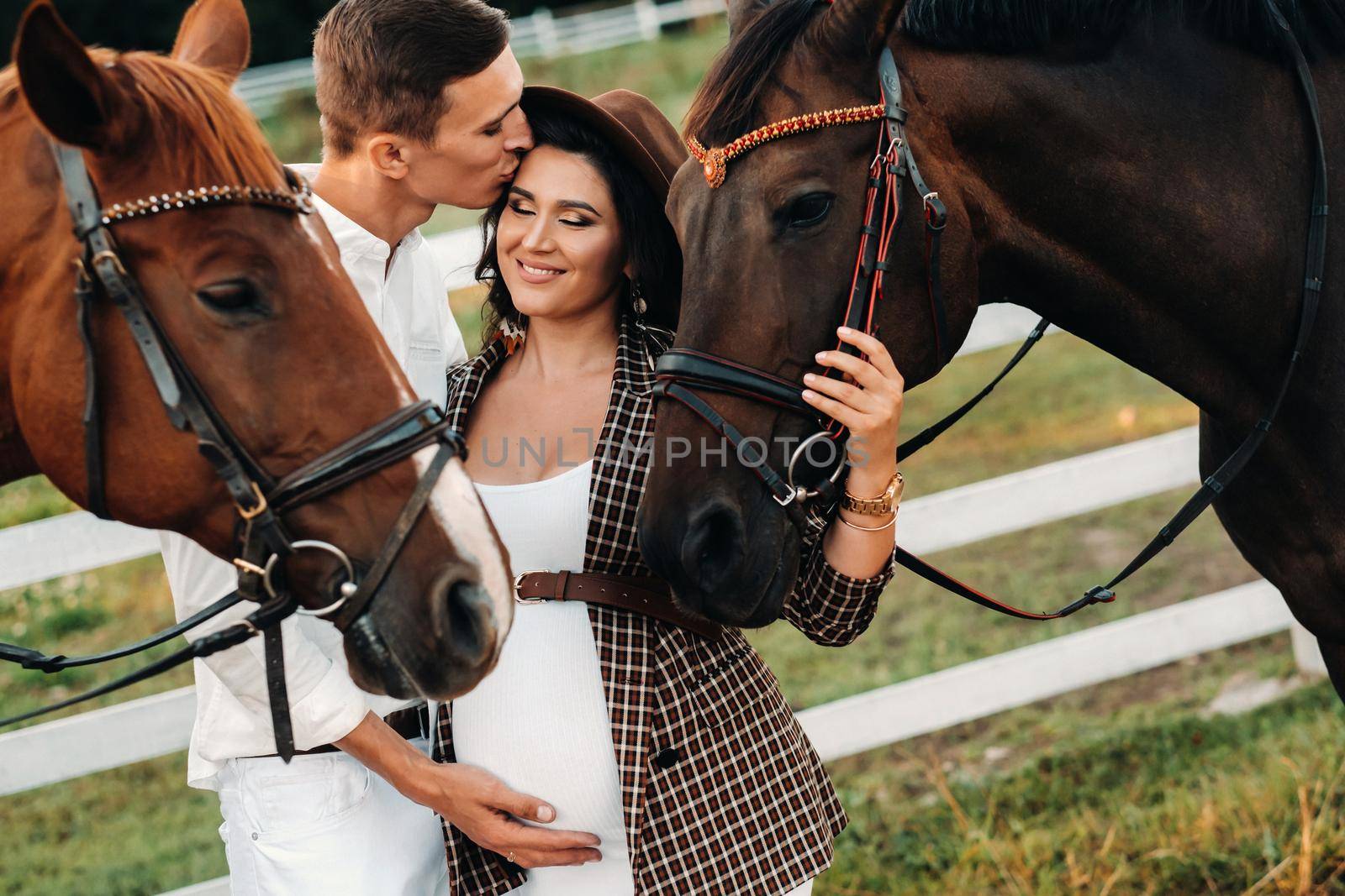 a pregnant girl in a hat and her husband in white clothes stand next to the horses near the horse corral.Stylish pregnant woman with a man with horses.Family by Lobachad