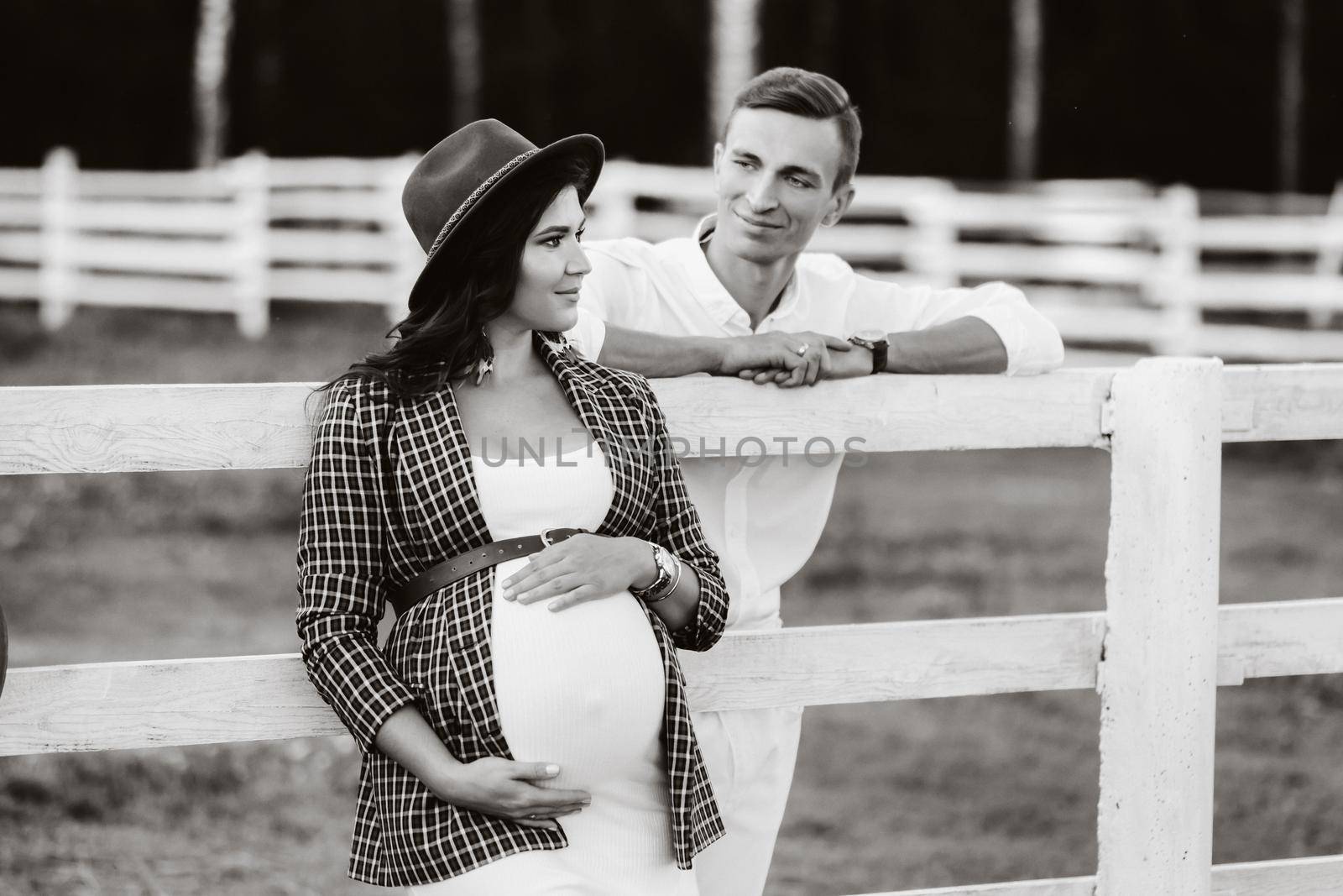 a pregnant girl in a hat and her husband in white clothes stand next to the horse corral.a stylish couple waiting for a child stand on the street near the horse corral.black and white photo by Lobachad