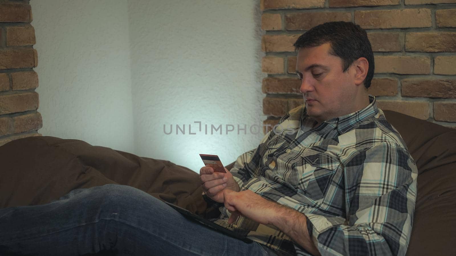 A man in a plaid shirt and jeans sitting on a chair bag in his hands holding a tablet and credit card makes online purchases, pay bills. Soft panorama