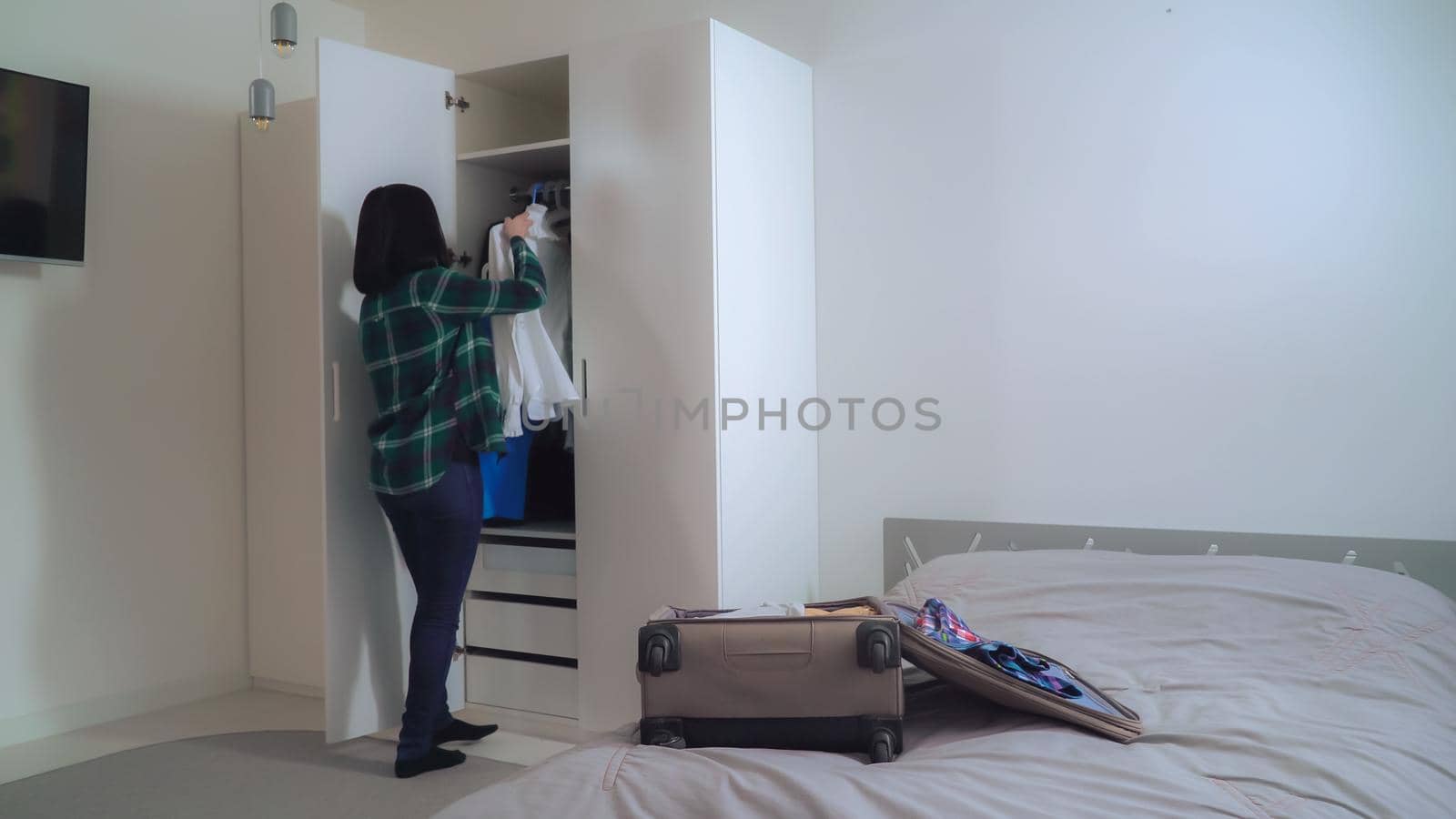 Young woman wearing in casual shirt and jeans at home. Brunette unpacks a suitcase in flat. Girl taking clothes on hangers and hanging in the closet in apartment.