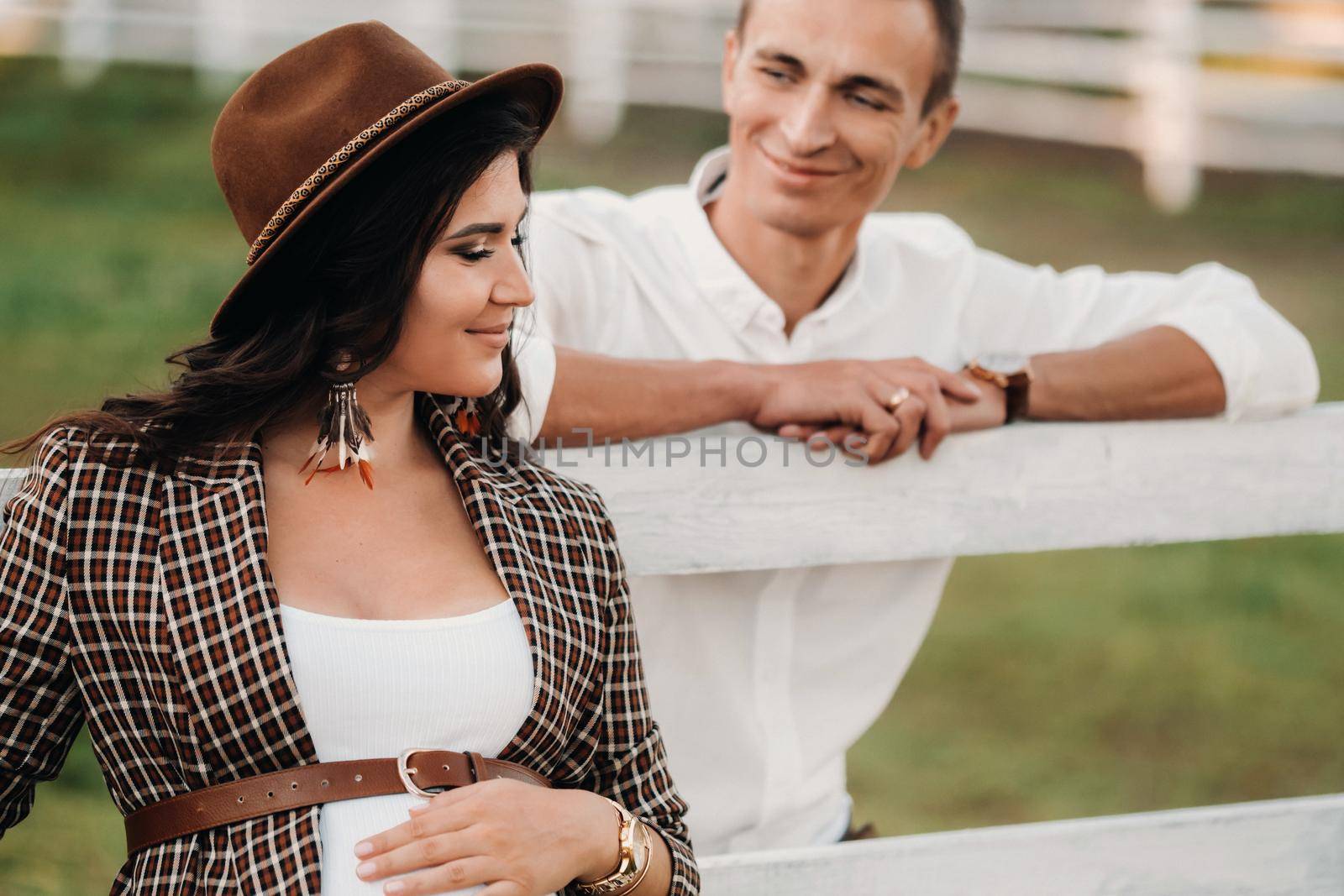 a pregnant girl in a hat and her husband in white clothes stand next to a horse corral at sunset.a stylish couple is waiting for a child in nature by Lobachad