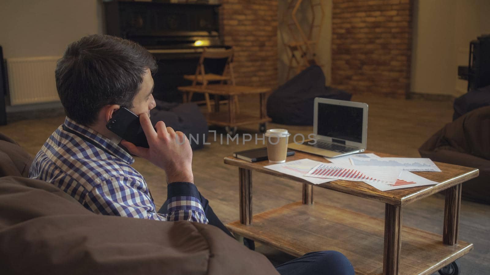 A man in shirt sitting in upholstered furniture, keep a schedule in hand and talking on the phone. On the table are the working paper and cost laptop.