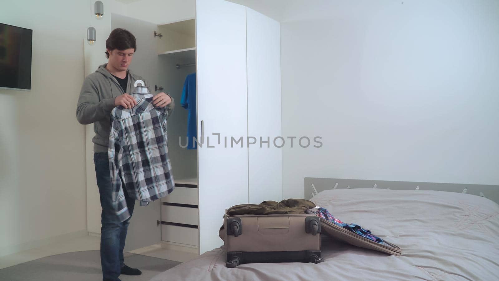 Young happy guy packing clothes in suitcase. Smiling casual businessman planning business trip in apartment with modern interior.