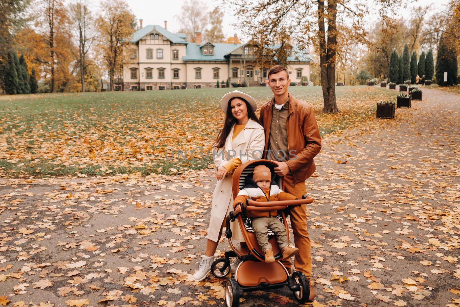 Father and mother on a walk with a child in a stroller in the autumn Park on the background of the estate. A family walks through the Golden autumn nature Park by Lobachad