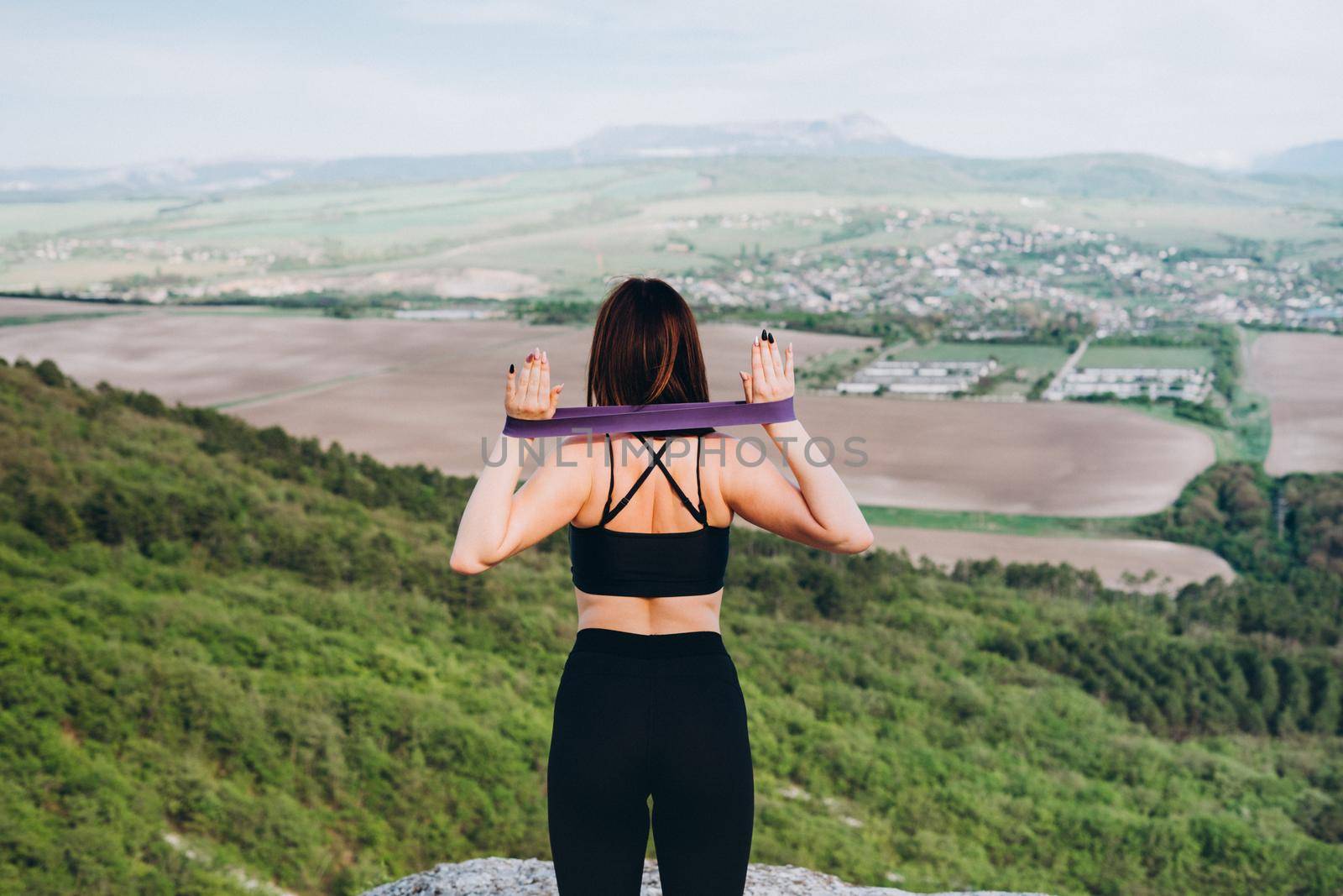 The girl stands with her back to the camera and trains with a rubber band in nature. The girl is engaged in sports in nature. Purple rubber band for stretching and swaping muscles of hands and feet.