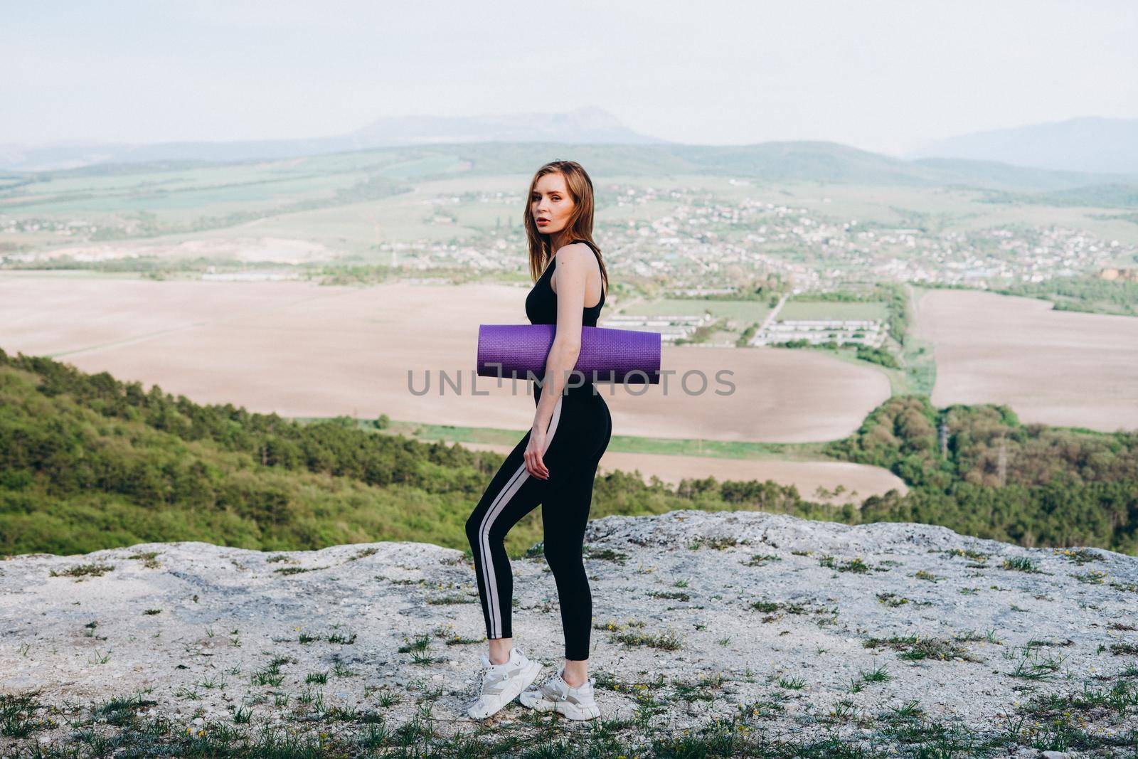 A girl athlete with a purple yoga mat under her armpit. A beautiful athlete in the mountains.